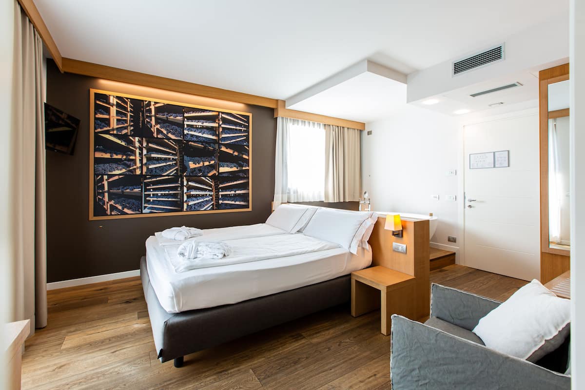 A modern hotel room with a large bed, a bedside table, a chair, and a large abstract artwork on the wall.