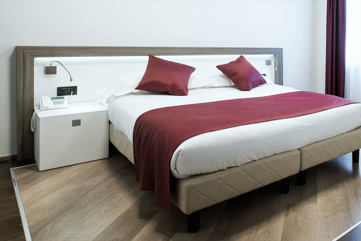 A neatly made bed with a white headboard, two red pillows, and a red throw blanket. There is a white bedside table with a lamp and a small device on it. The room has wooden flooring and a plain white wall.