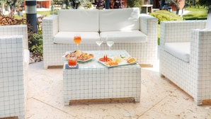 An outdoor seating area with a white sofa and matching chairs, a tiled coffee table with food and drinks, and a pergola overhead. - Hotel Corte del Paggio (Valeggio sul Mincio)