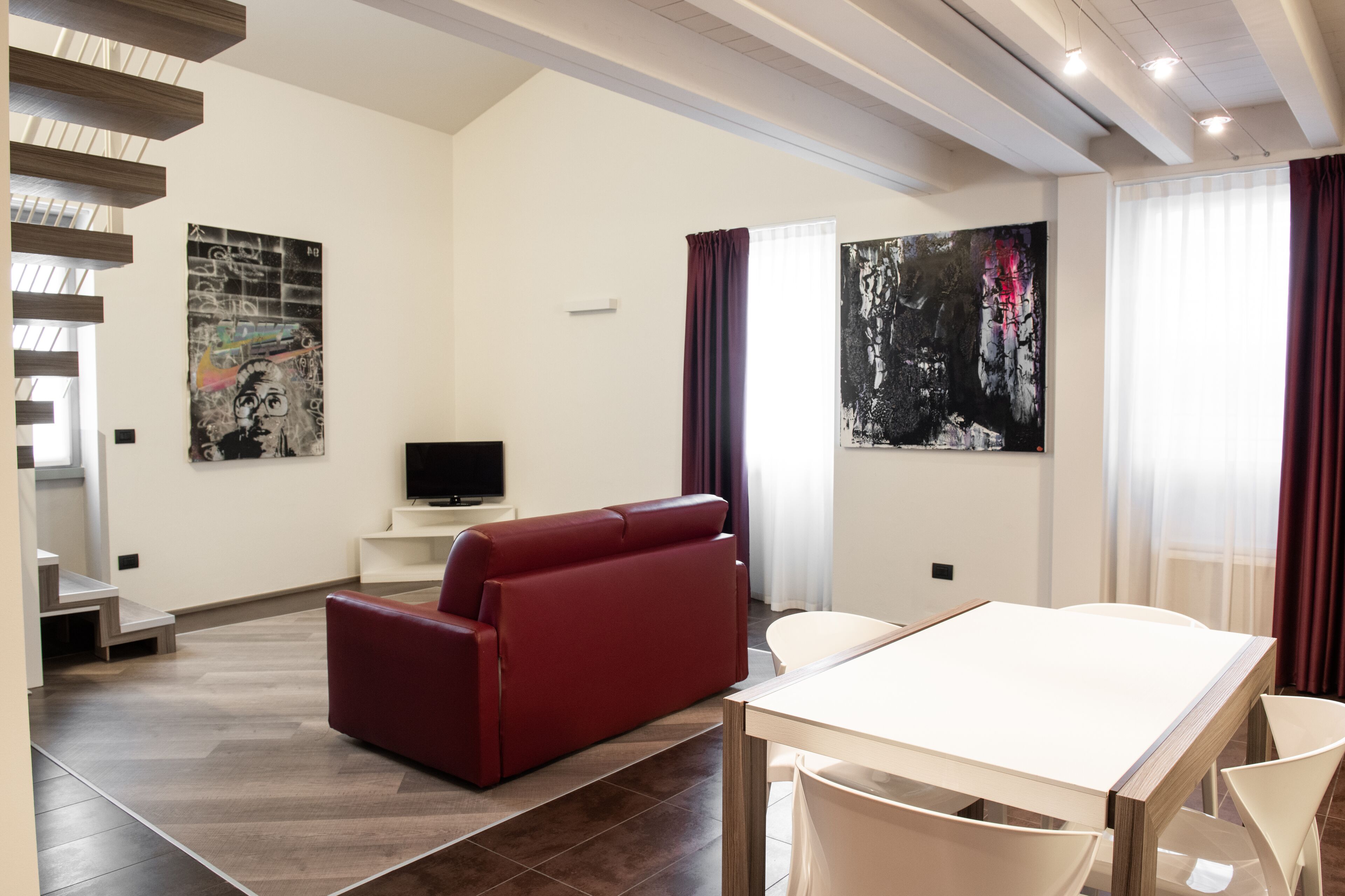 Uma sala de estar moderna com um sofá vermelho, uma mesa branca e duas cadeiras brancas. Há uma televisão de tela plana sobre um suporte branco. O cômodo possui uma escada com degraus de madeira e um corrimão branco.