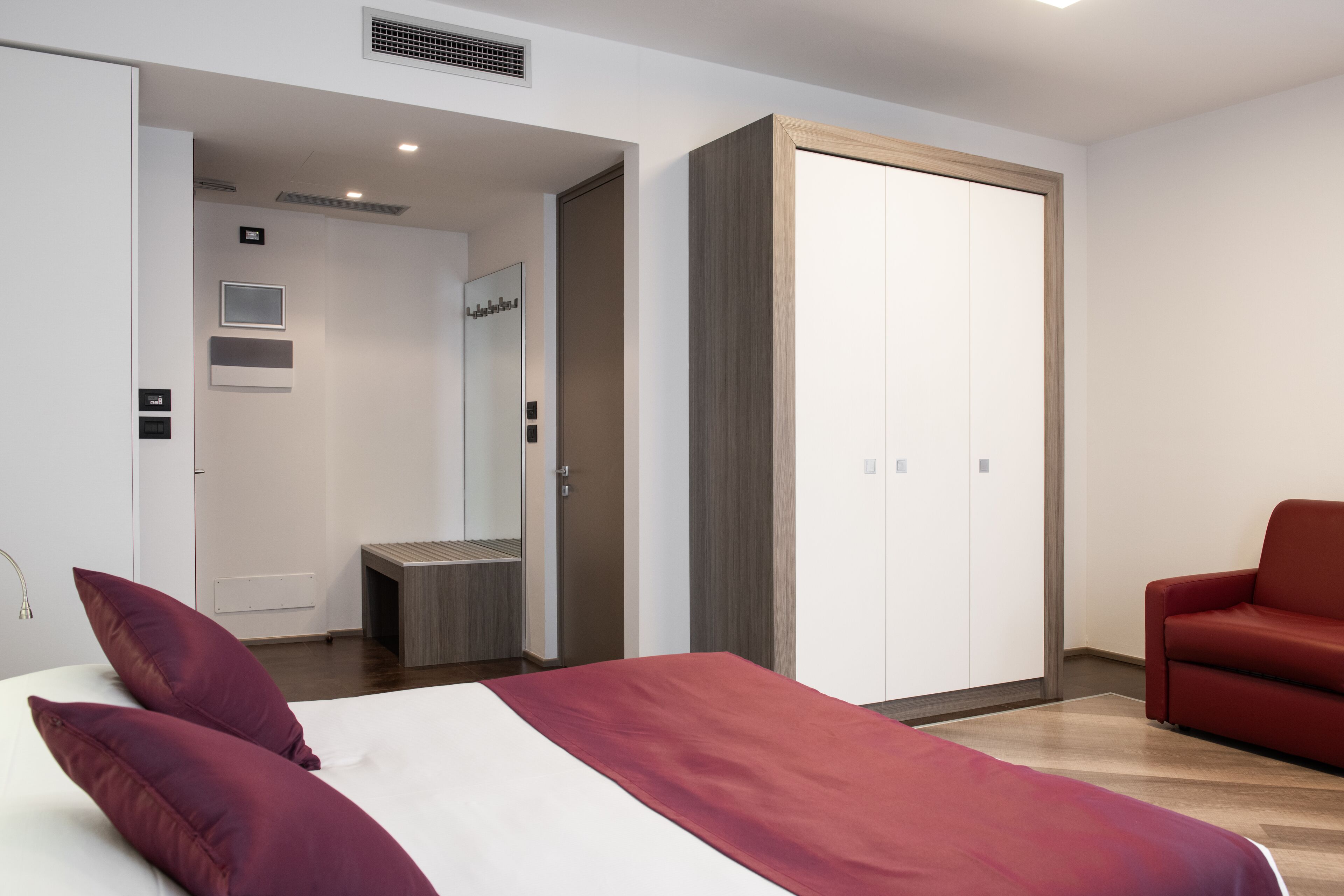 Une chambre d’hôtel moderne avec un lit, un canapé rouge, une armoire et une salle de bain visible à travers une porte ouverte.