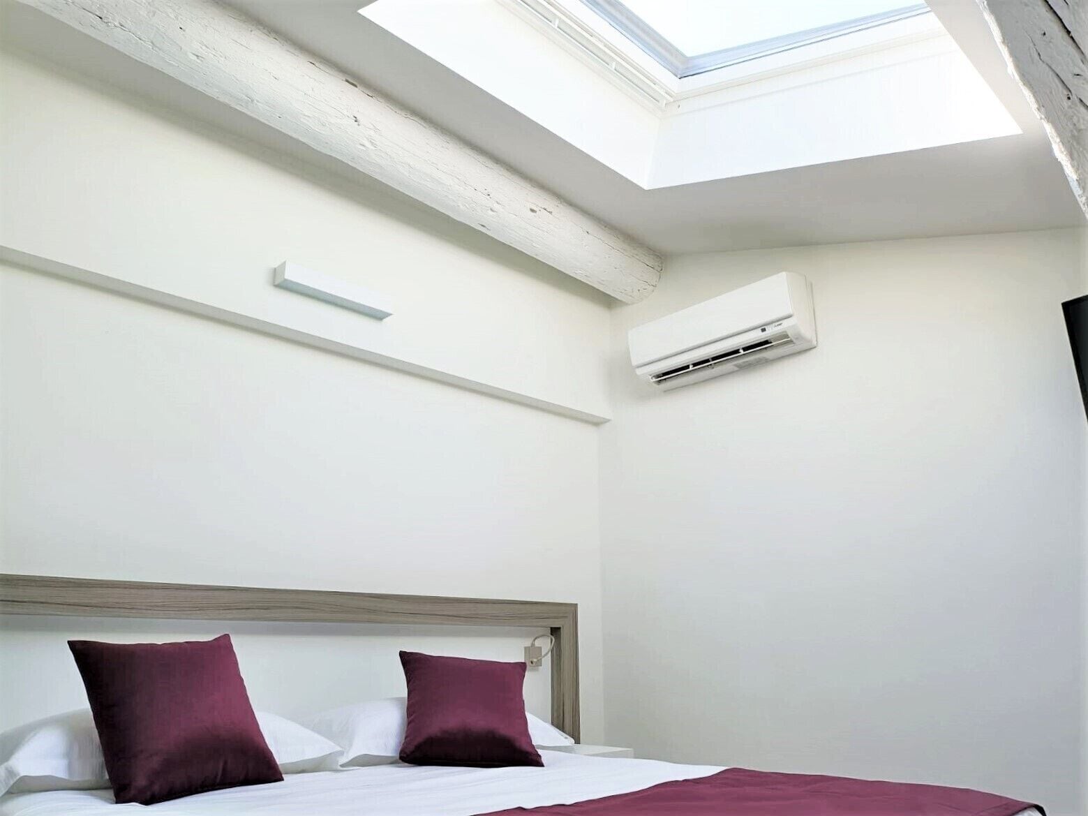 Ein Hotelzimmer mit Dachfenster, einem Bett mit weißer und purpurroter Bettwäsche sowie einer weißen Klimaanlage.