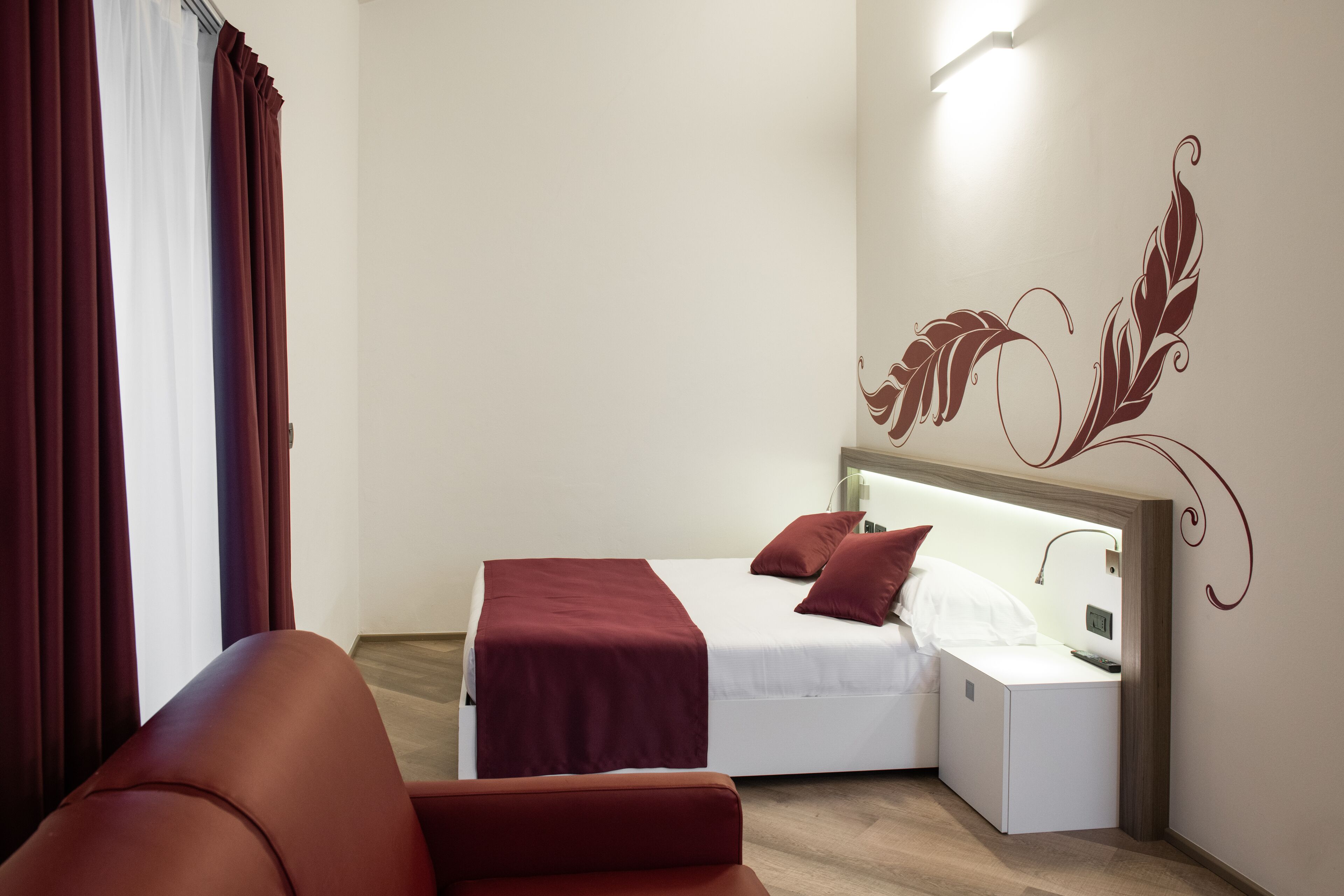 A compact hotel room with a bed, a nightstand, a red chair, and a decorative wall design.