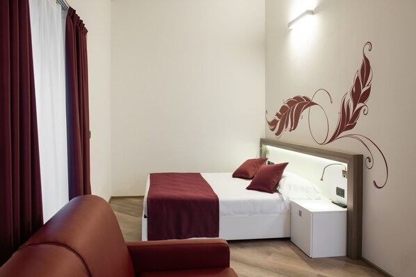 A compact hotel room with a bed, a nightstand, a red chair, and a decorative wall design.