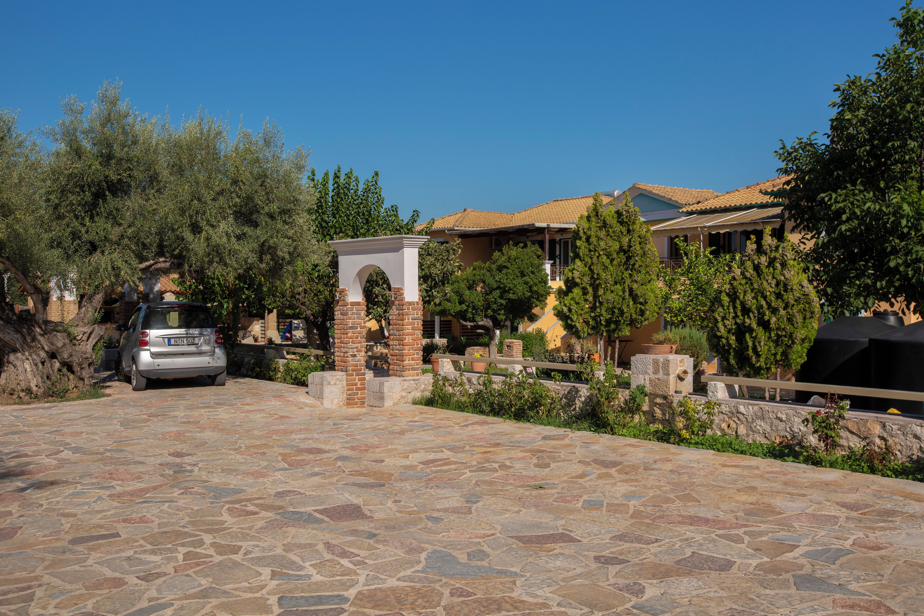 A stone-paved driveway leading to a house with a covered patio area, a barbecue grill, and a car parked nearby.