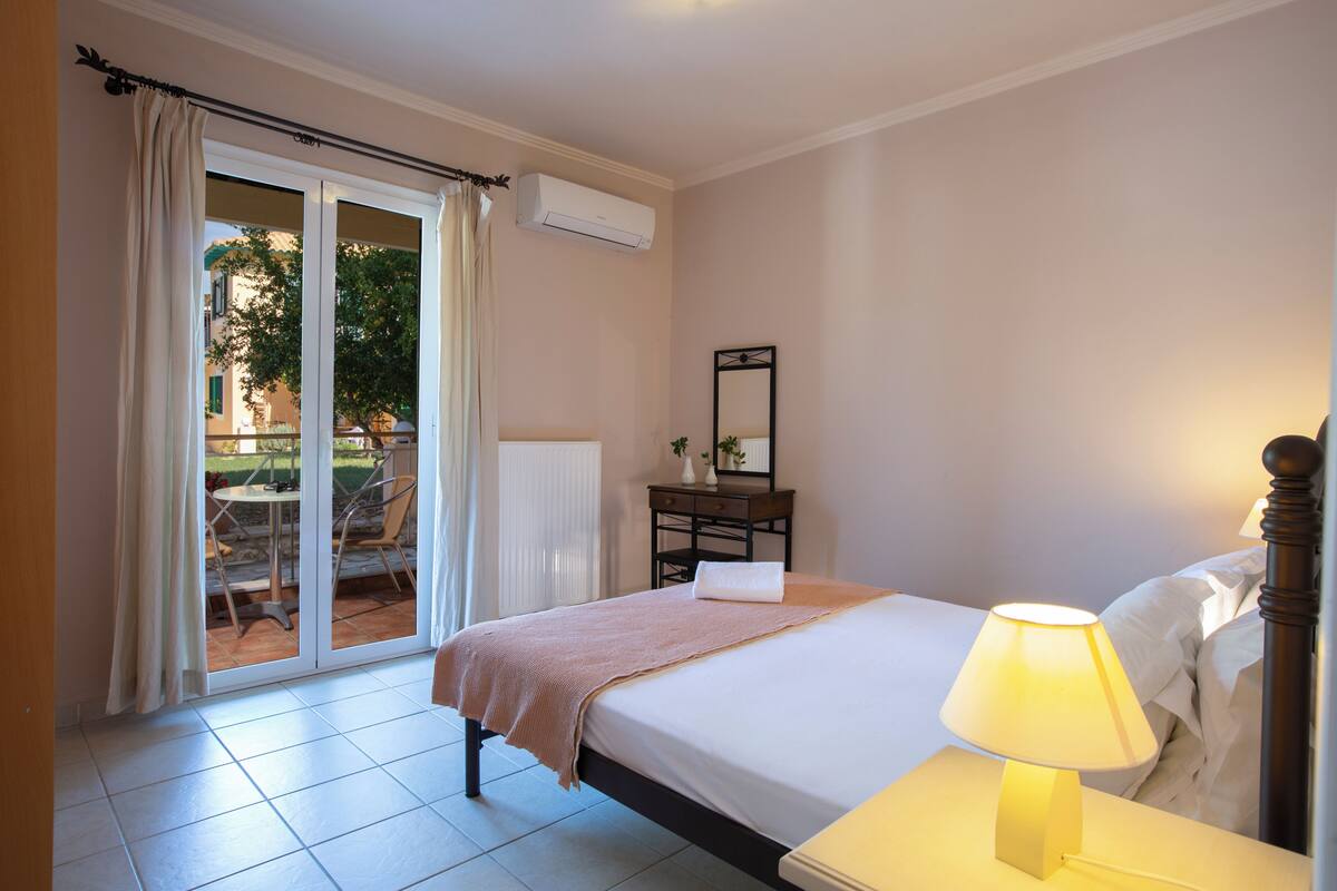 Ein Hotelzimmer mit einem Bett, einem Nachttisch mit Lampe, einem Schminktisch, einem Spiegel und einer Glastür, die zu einem Außenbereich mit Tisch und Stühlen führt.