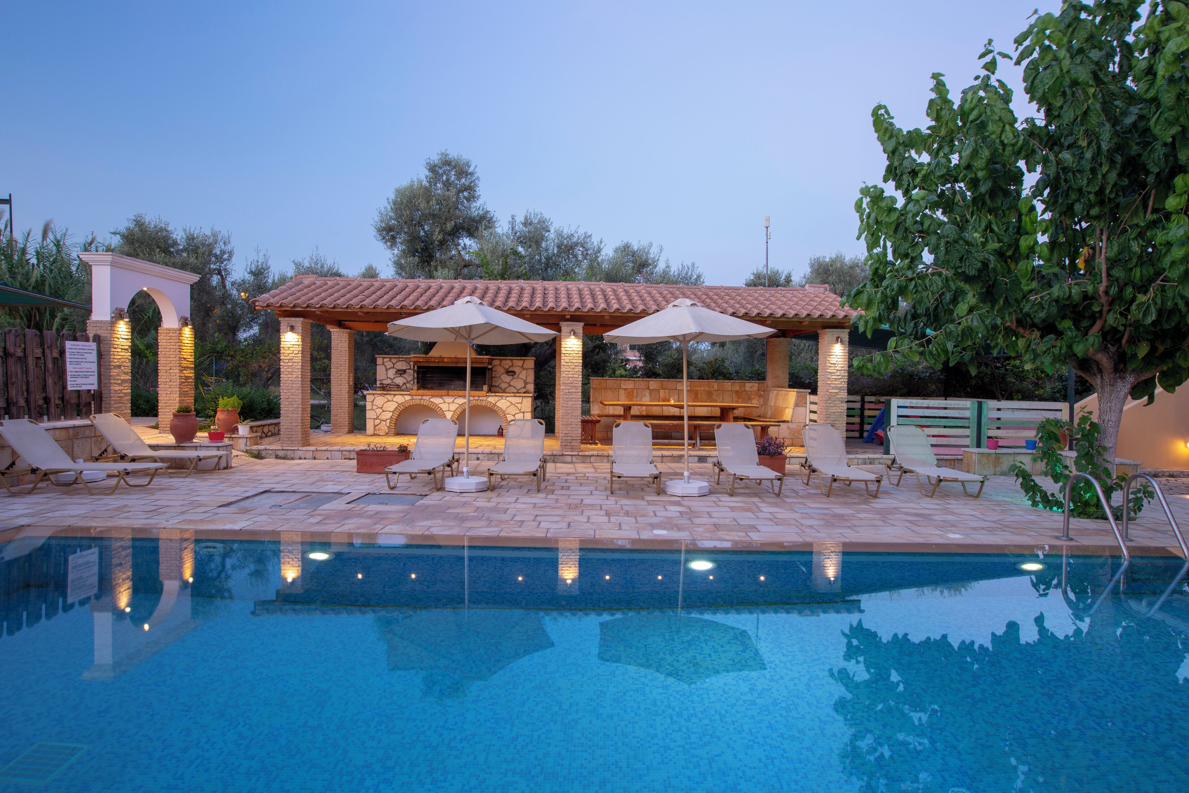 Un área junto a la piscina con sillones de descanso, una zona de barbacoa y una pérgola con sombrillas.