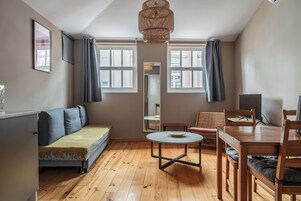A room with a sofa, a round table, a dining table with chairs, a mirror, and two windows with curtains.