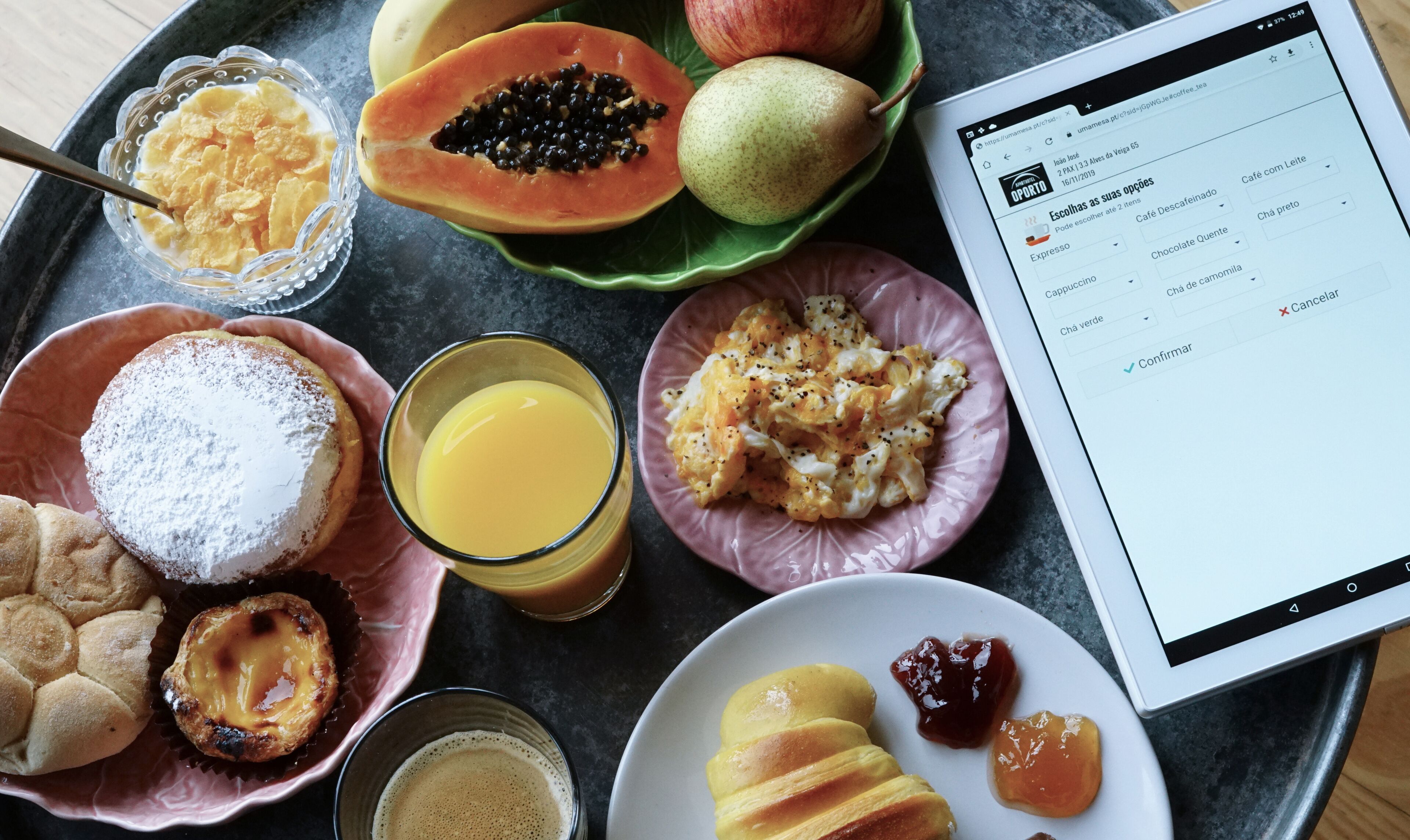 Bandeja de desayuno con diversos alimentos, incluyendo pasteles, un vaso de jugo de naranja y una tableta mostrando un menú.