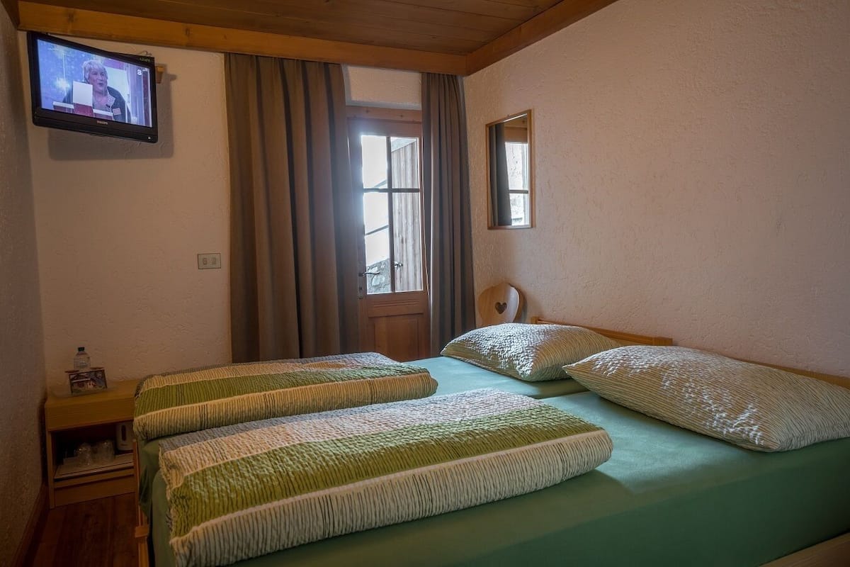 A room with two beds, a television mounted on the wall, a wooden headboard, and a window with curtains.