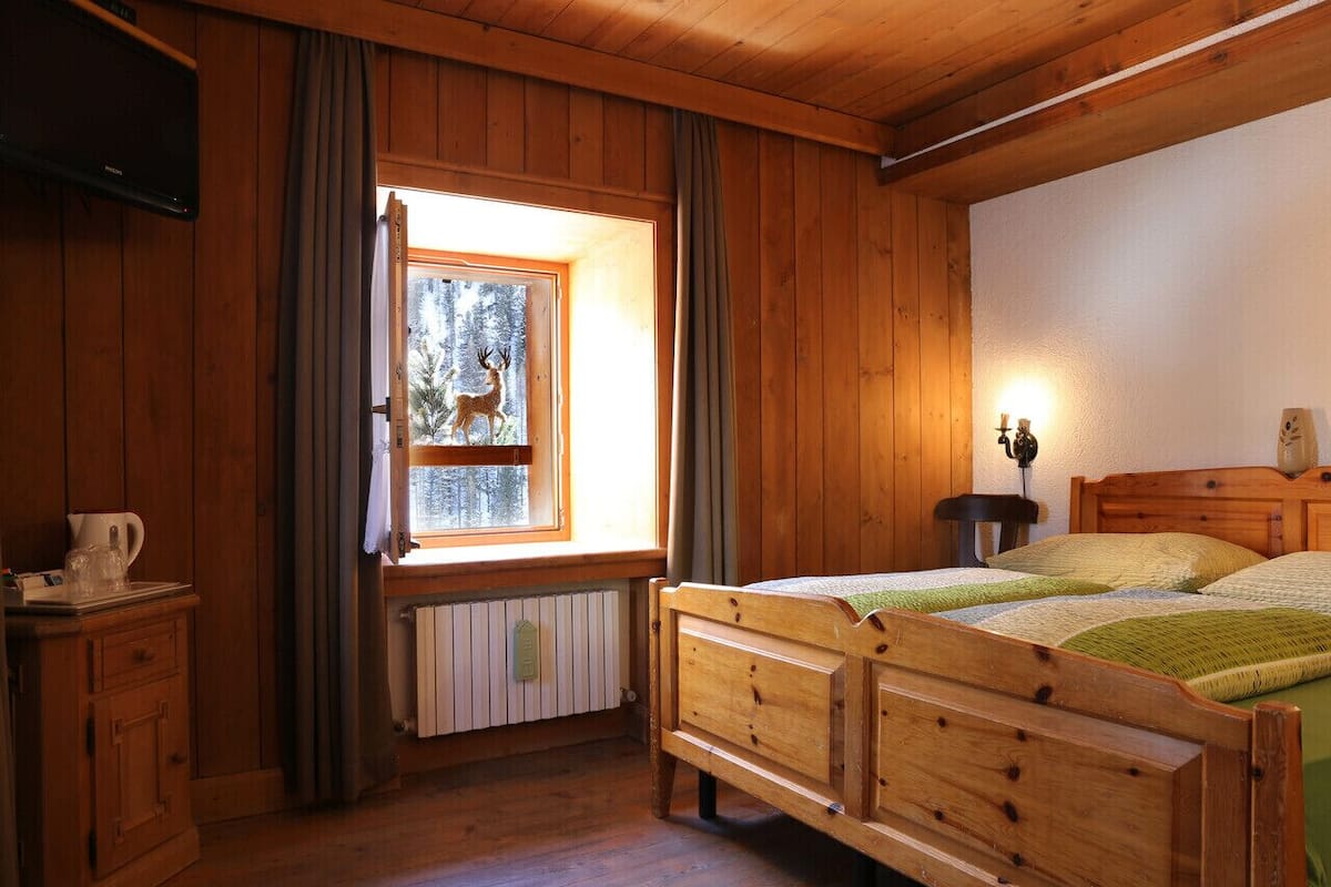 A wooden cabin room with a bed, a window with curtains, a radiator, and a television on the wall.