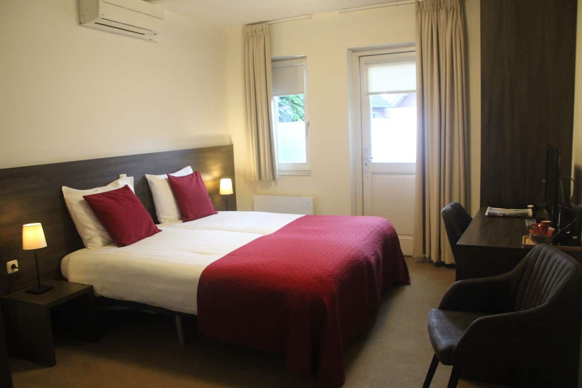 A hotel room with a bed, two bedside lamps, a desk with a chair, and a window with curtains.