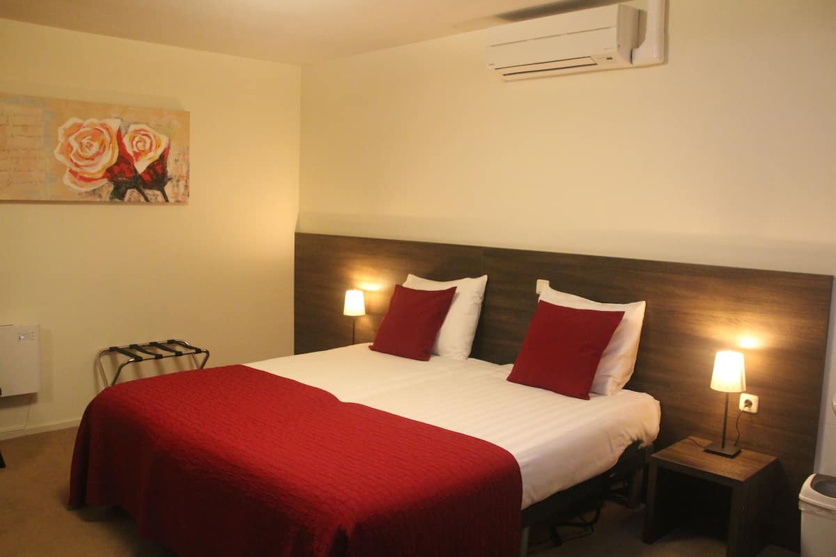 A hotel room with a large bed, red and white bedding, two bedside lamps, a wall-mounted air conditioner, and a painting of roses.