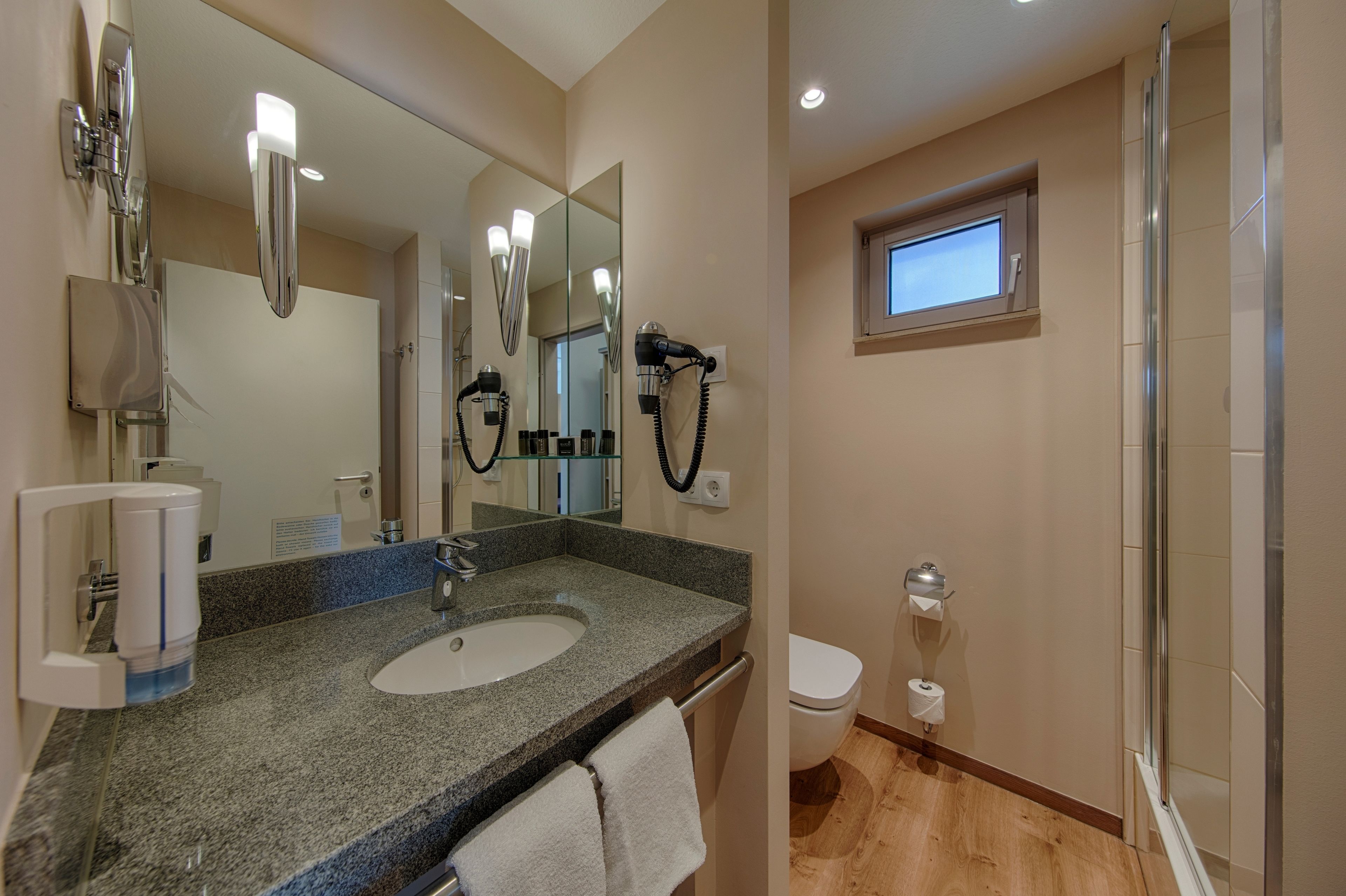 A bathroom with a granite countertop sink, a large mirror, a hairdryer, and a window above the toilet.
