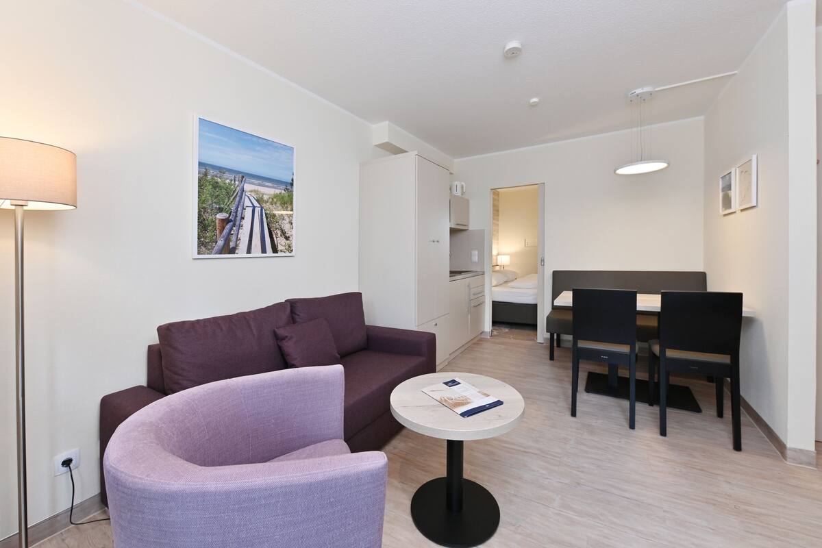 A modern hotel room with a purple armchair, a small round table, a dining area with a table and chairs, and a painting of a beach on the wall.