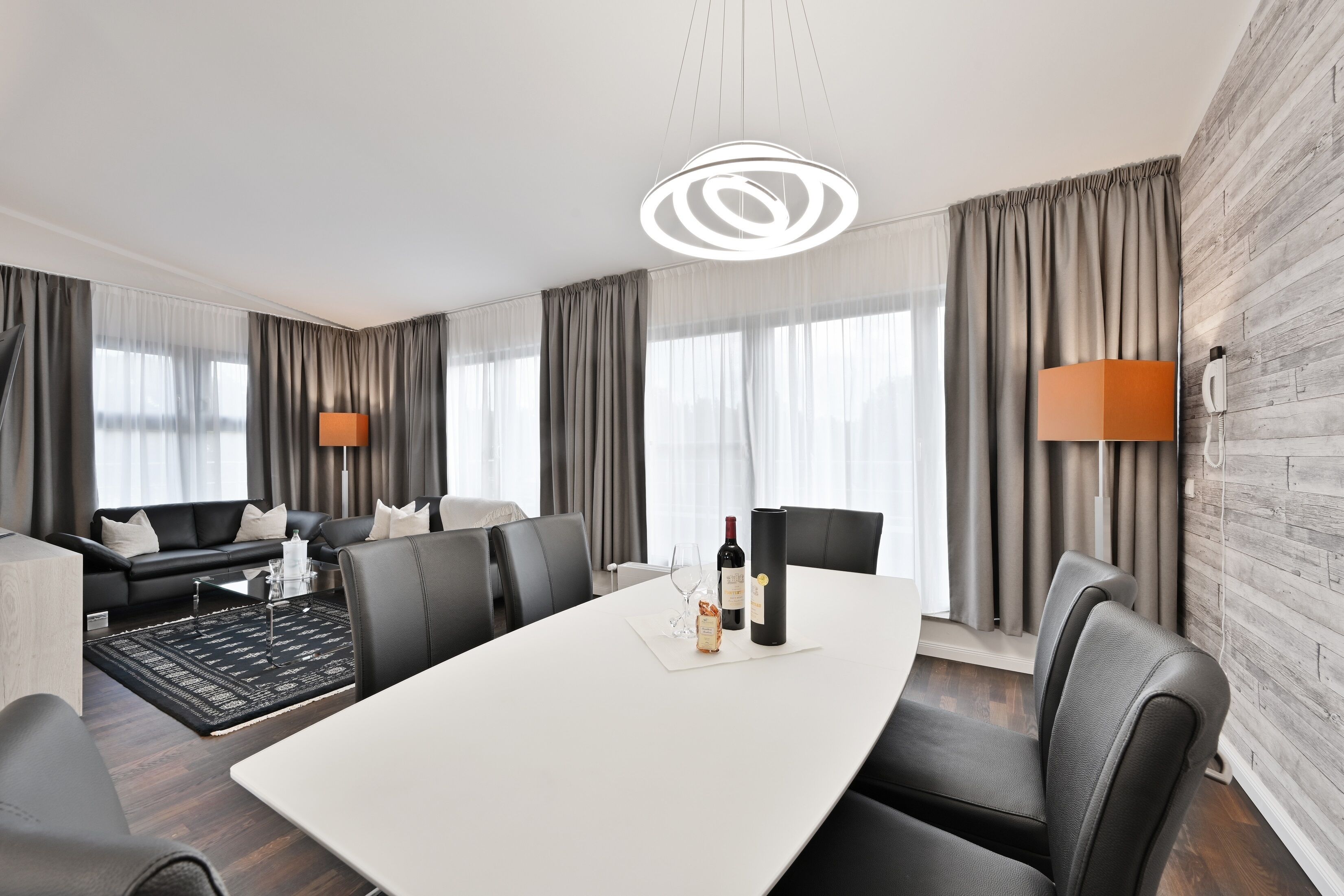 A modern hotel room with a large dining table, leather chairs, a sofa, and a large window with curtains.