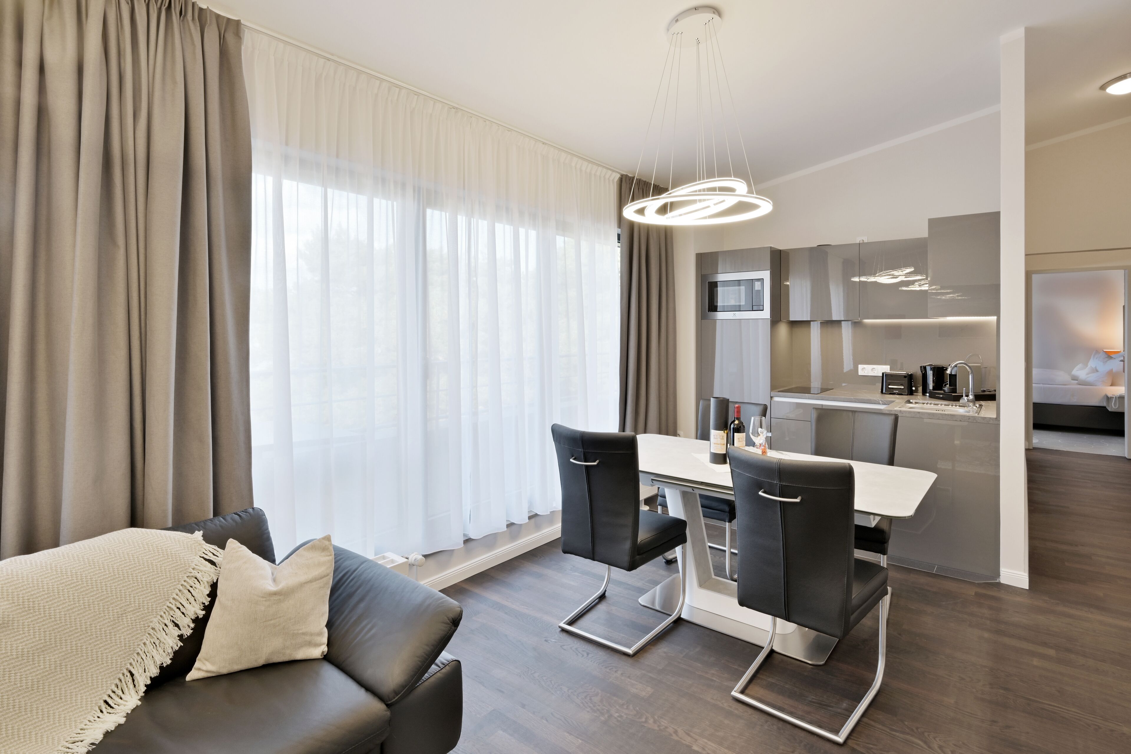 A modern living room with a sofa, dining table, and chairs, a kitchen area with a microwave, and a large window with curtains.