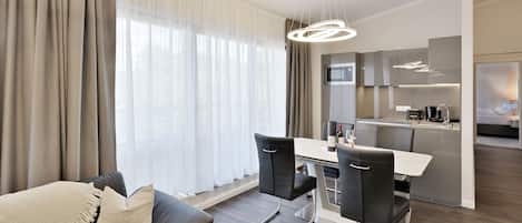 A modern living room with a sofa, dining table, and chairs, a kitchen area with a microwave, and a large window with curtains.