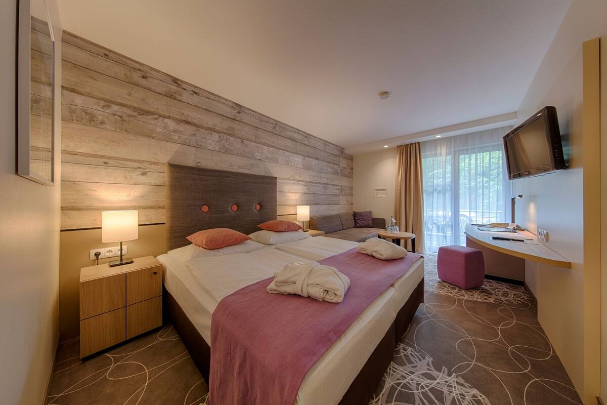 A hotel room with a large bed, bedside tables, a desk, a television, and a window with curtains.