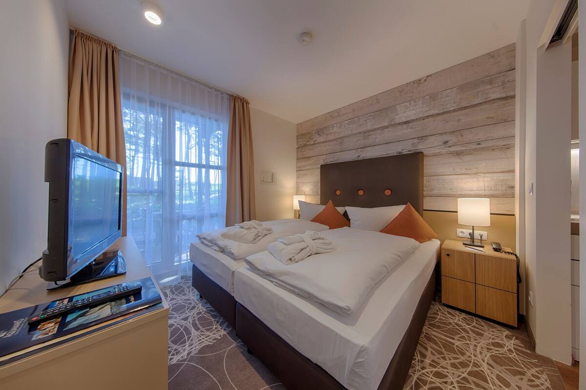 A hotel room with a large bed, a TV, a bedside table with a lamp, and a view of trees through the window.