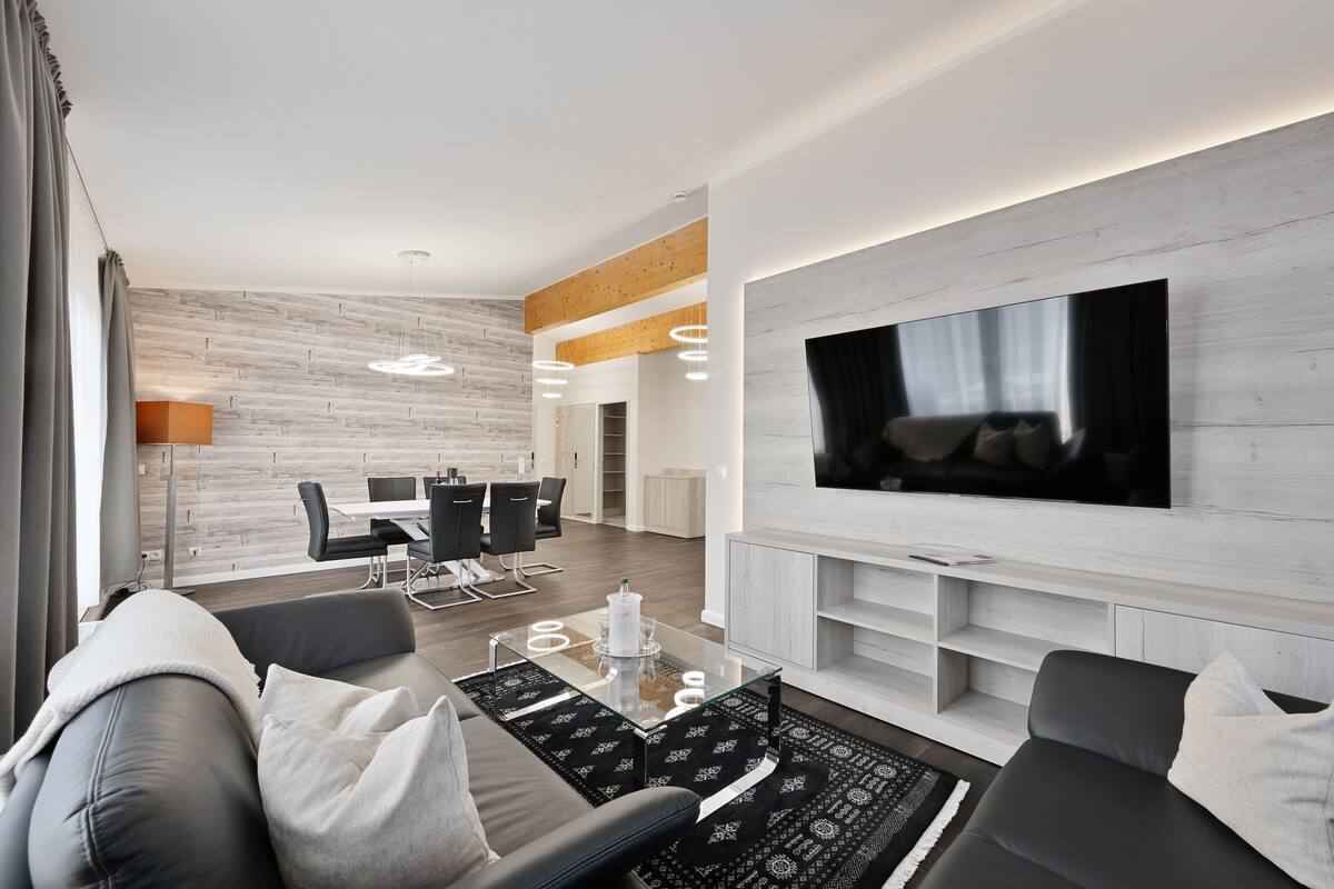 A modern living room with a large flat-screen TV mounted on the wall, a grey sofa with cushions, a glass coffee table, and a dining area with a white table and chairs.