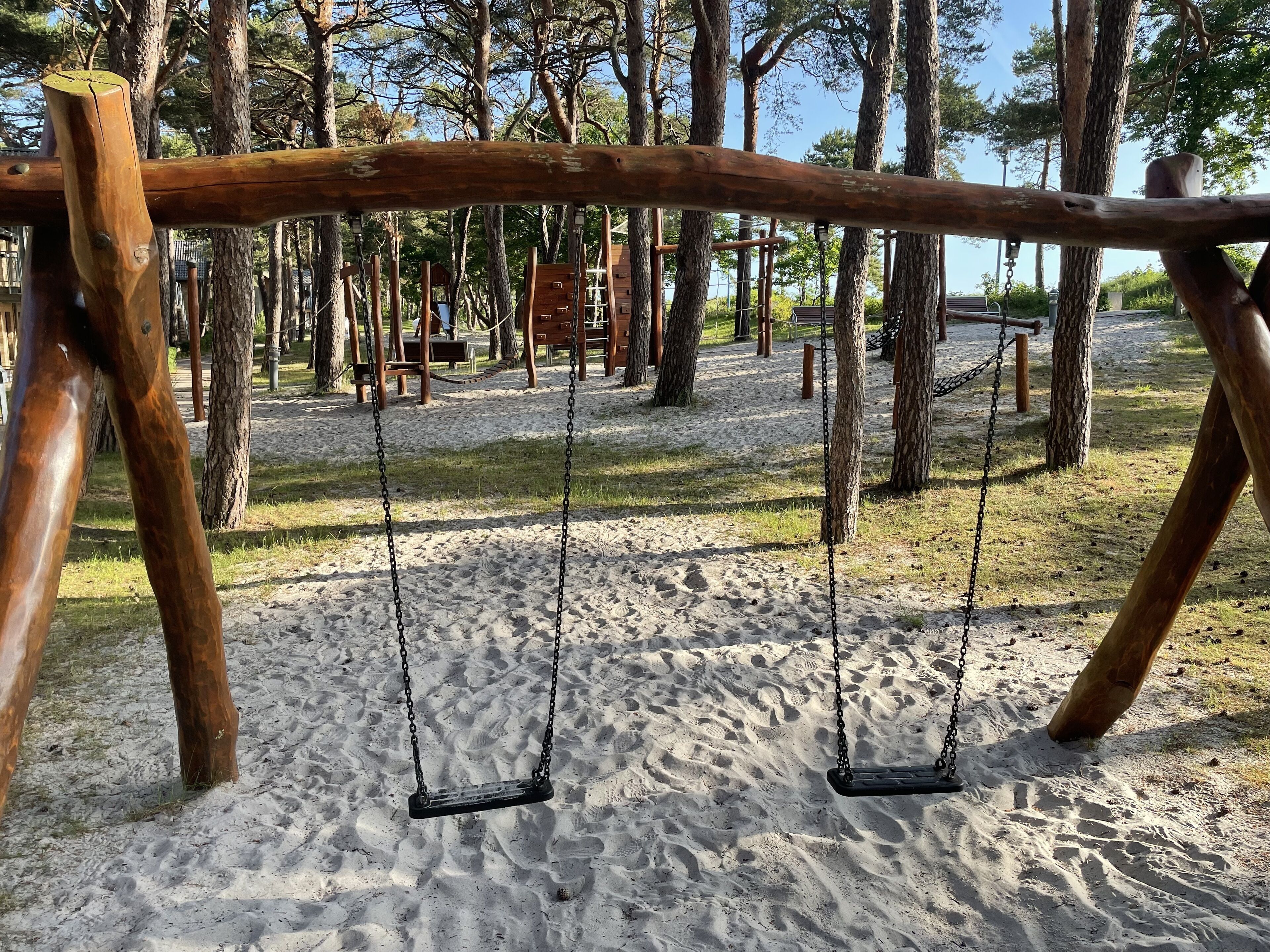 A wooden swing set in a park with sand ground and trees in the background.