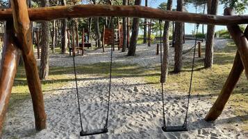 A wooden swing set in a park with sand ground and trees in the background.