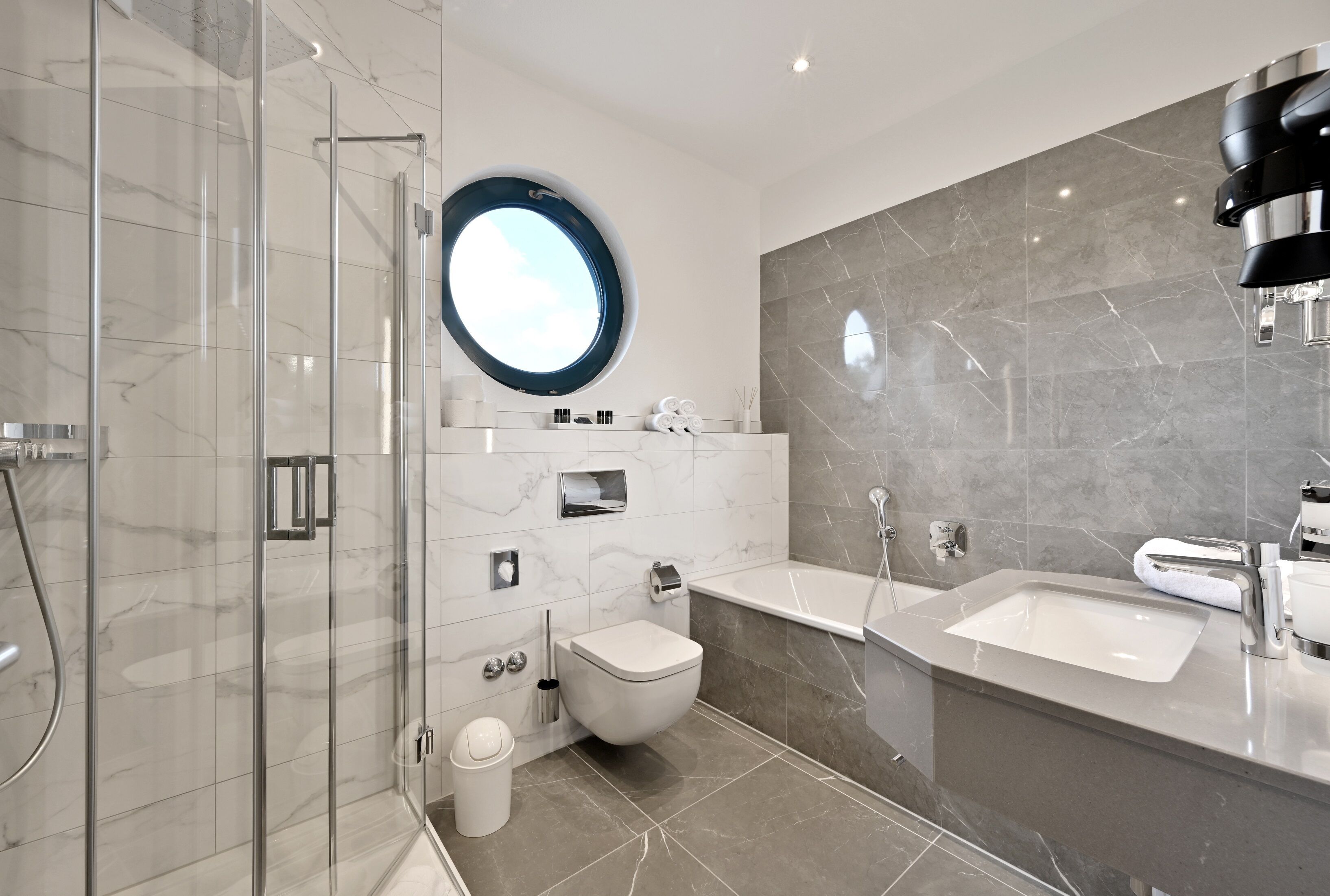 Un baño moderno con ducha envuelta en vidrio, espejo redondo, inodoro y dos lavamanos.