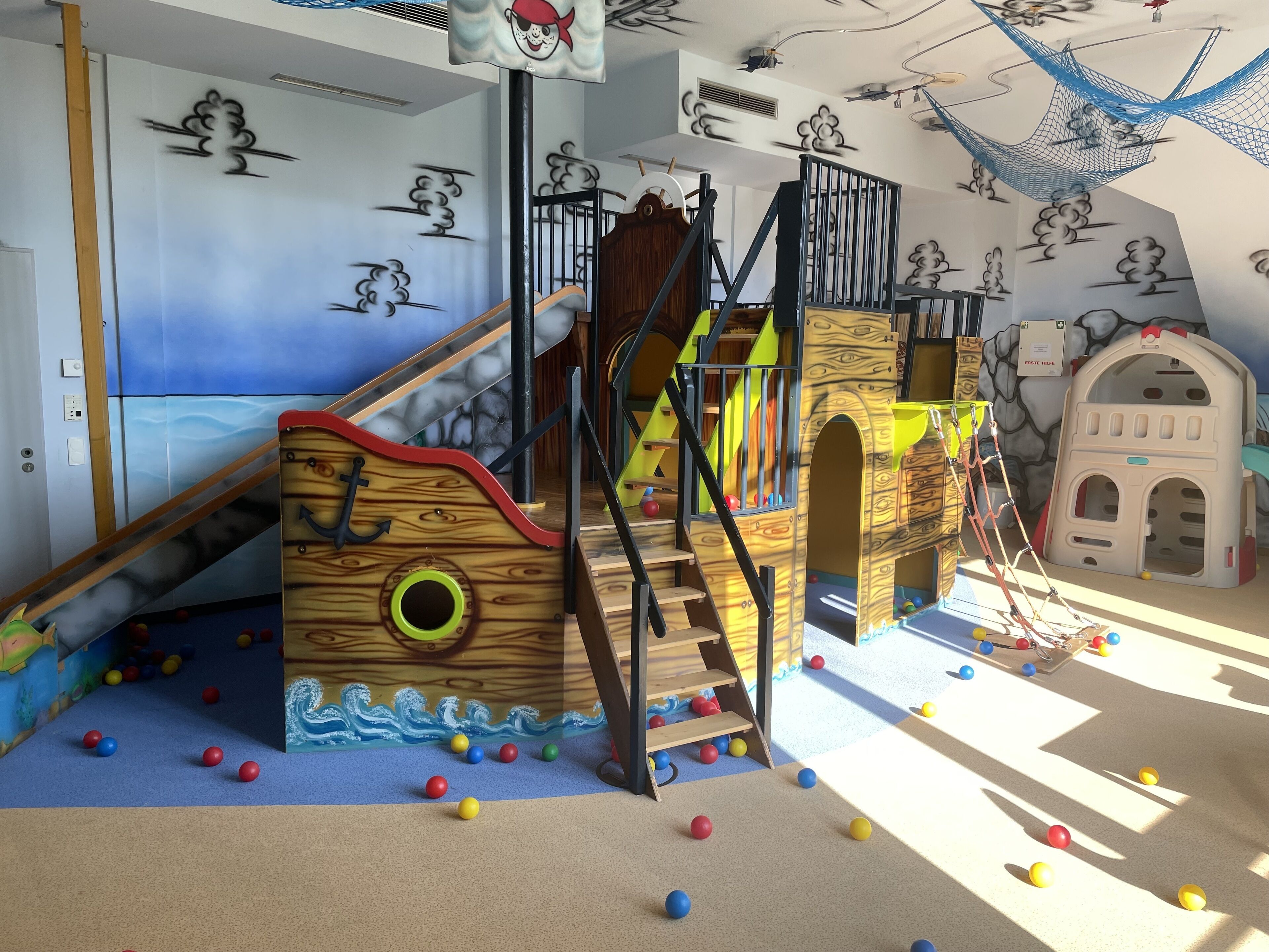 Ein Spielbereich mit einer hölzernen Spielanlage im Schiffdesign, einer Rutsche und einem kleinen Haus-ähnlichen Gebäude.