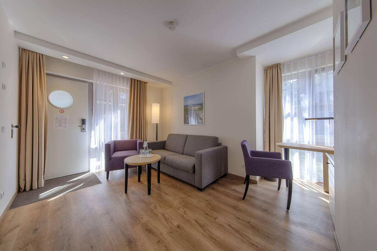 A modern hotel room with a sofa, a chair, a small table, and a window with curtains.