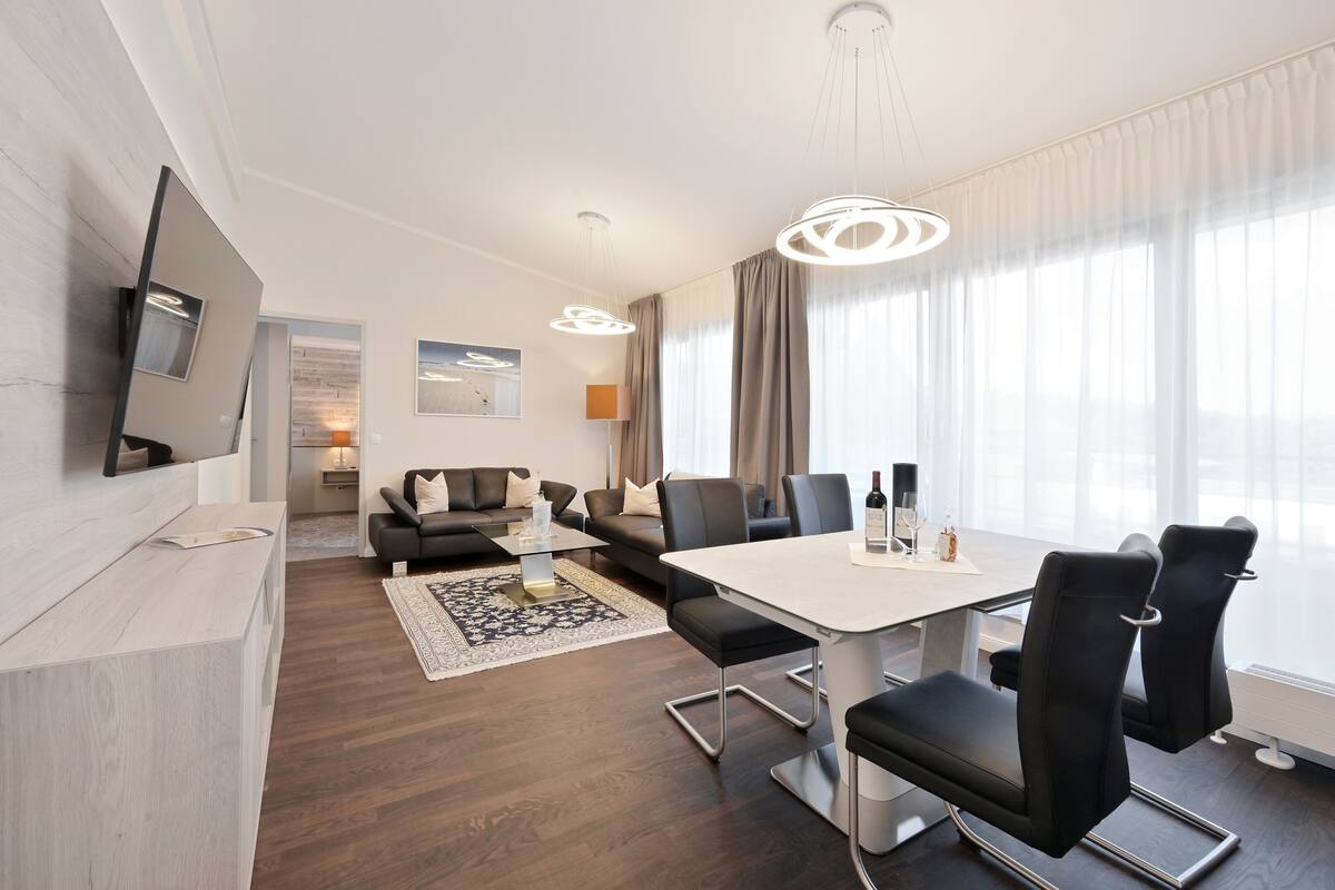 A modern hotel room with a dining area, a sofa, a TV, and a large window with curtains.