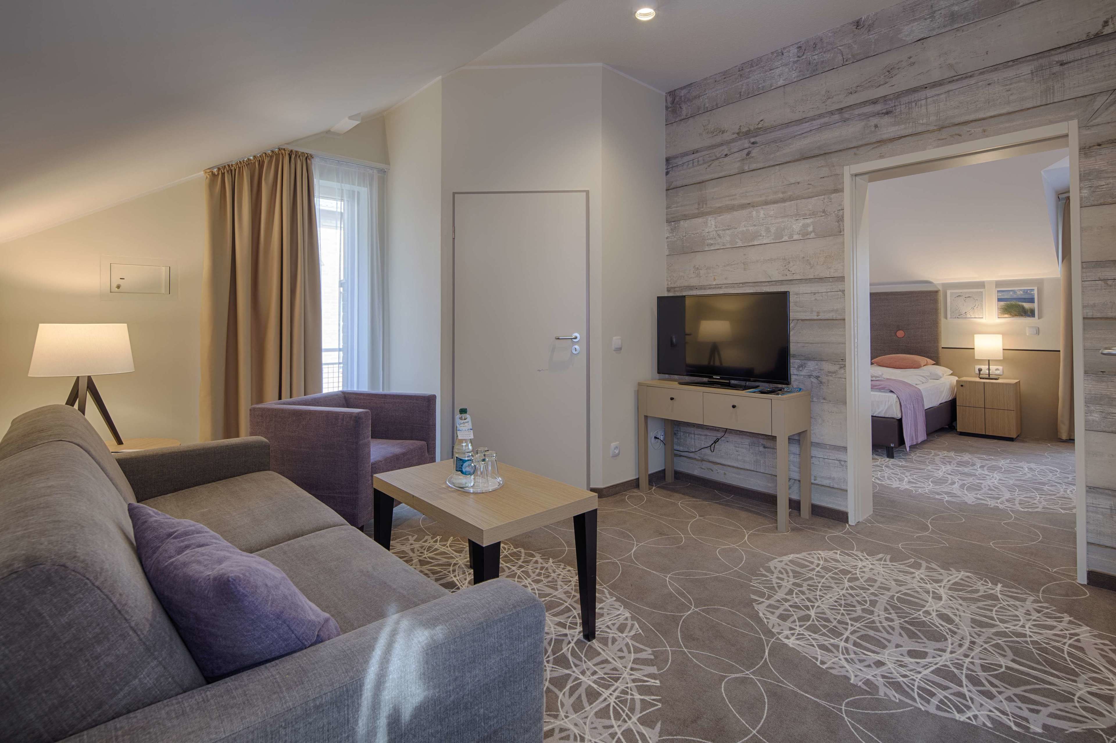 Een hotelkamer met een bank, een televisie, een kleintje tafeltje met een fles, een spiegel en een bed met nachtkastje.