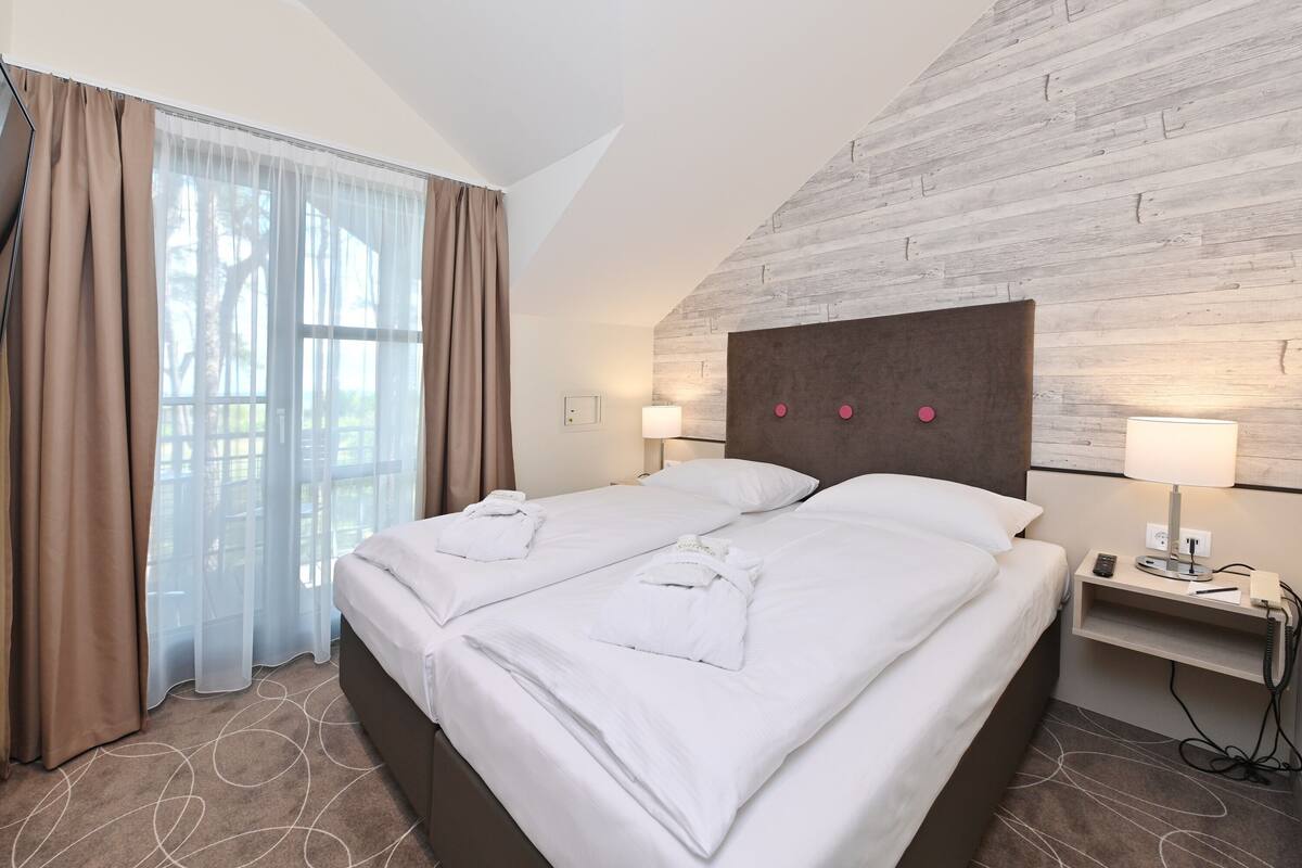 A hotel room with a large bed, two bedside tables, a headboard, and a window with curtains.