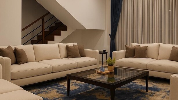 Un salon moderne avec deux canapés beige, une table basse en verre et un tapis à motifs bleus. On aperçoit un escalier au fond.