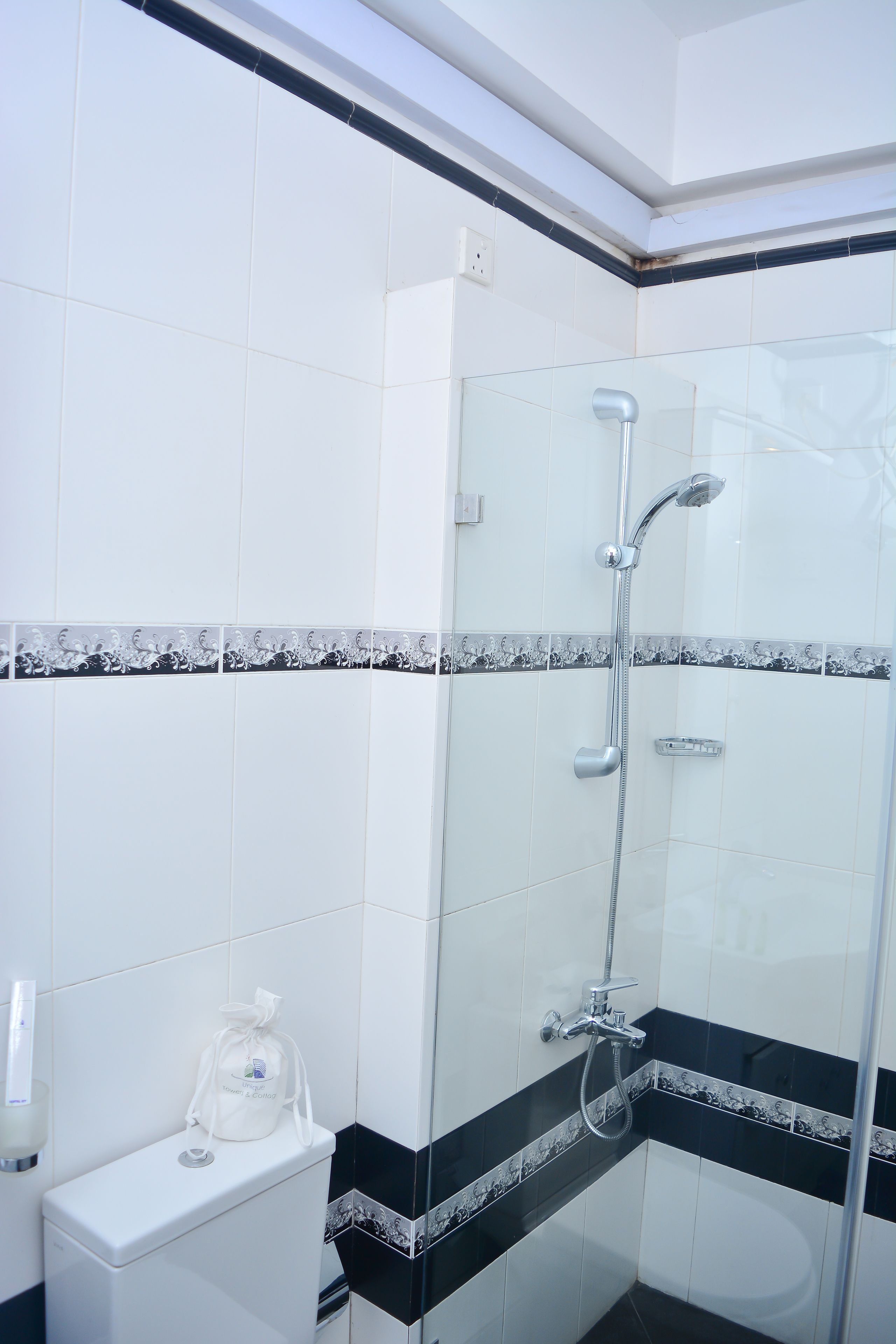 A bathroom with a glass shower enclosure, white tiles, and a black and white patterned border.