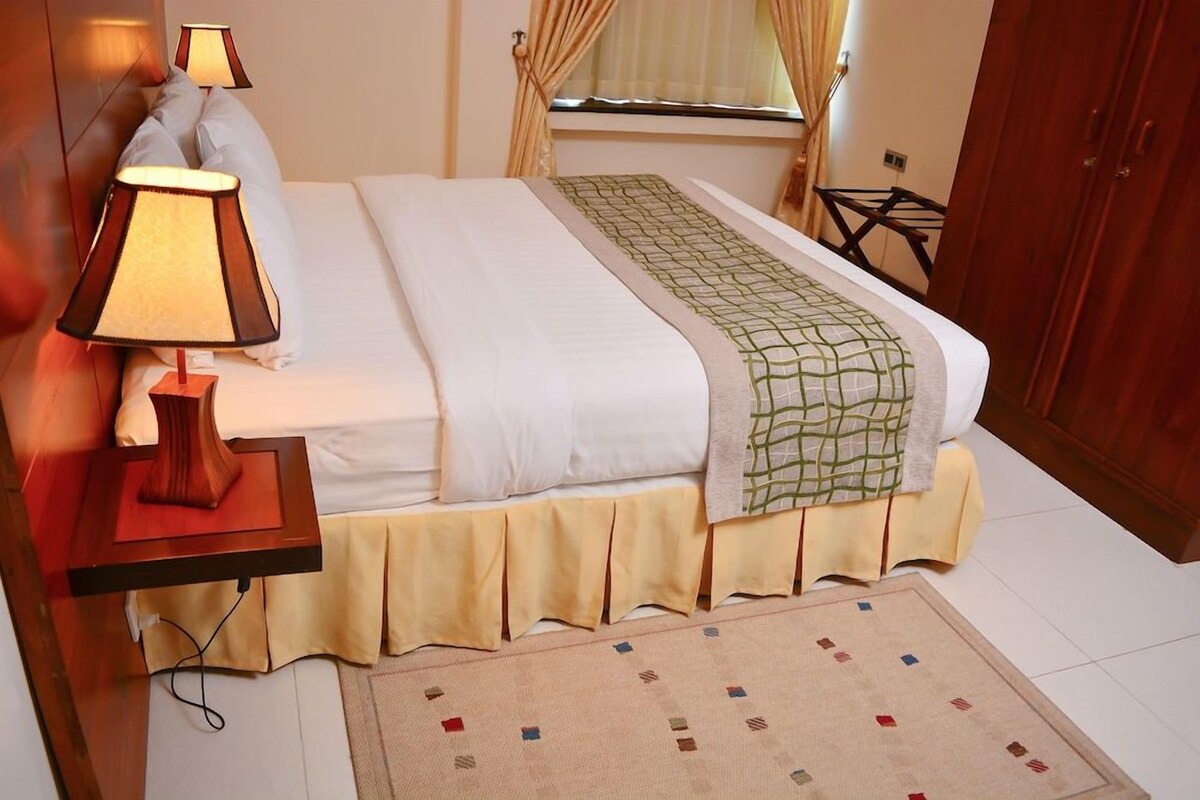 A hotel room with a bed, bedside lamps, a wooden nightstand, a window with curtains, and a small table with a lamp.