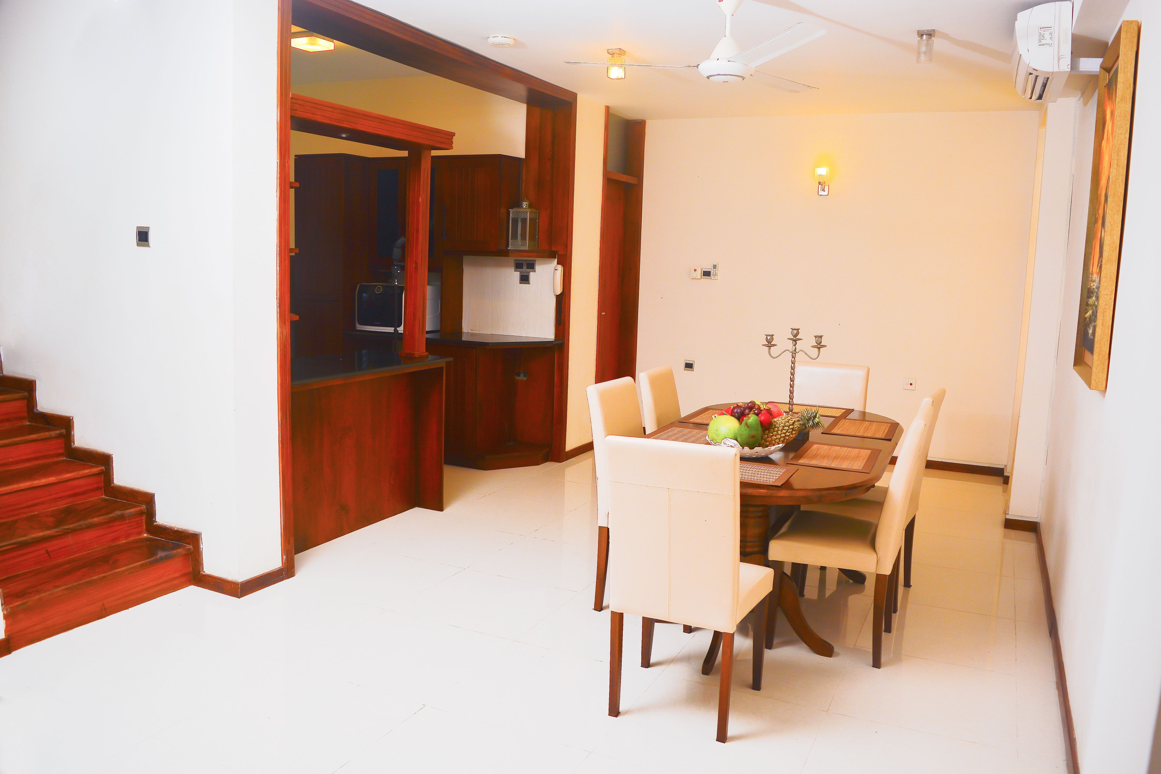 A dining area with a wooden table, white chairs, and a fruit bowl. There is a staircase leading to a kitchen area with wooden cabinets and a refrigerator.