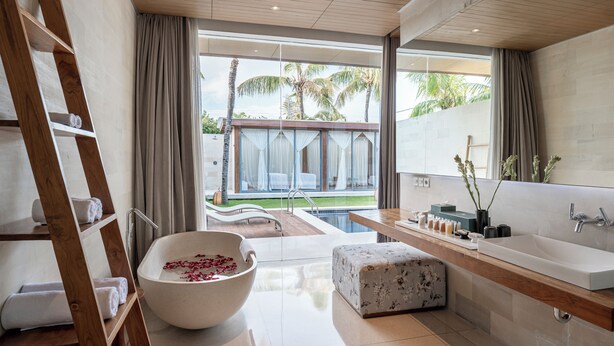 A modern bathroom with a freestanding bathtub, a wooden ladder, a large mirror, and a view of the pool and palm trees. - One Eleven Retreat (Keroboken Kelod)