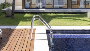 A modern outdoor pool area with a wooden deck, a glass-enclosed lounge with white curtains, and palm trees in the background. - One Eleven Retreat (Keroboken Kelod)