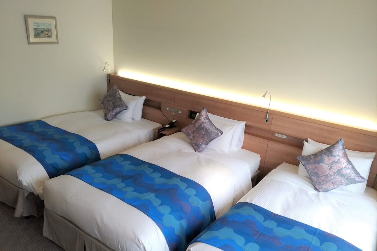 Habitación de hotel con tres camas, cada una con una colcha azul estampada y almohadas blancas. Tiene un cabecero de madera con iluminación integrada, una mesita de noche con teléfono y un cuadro en la pared.