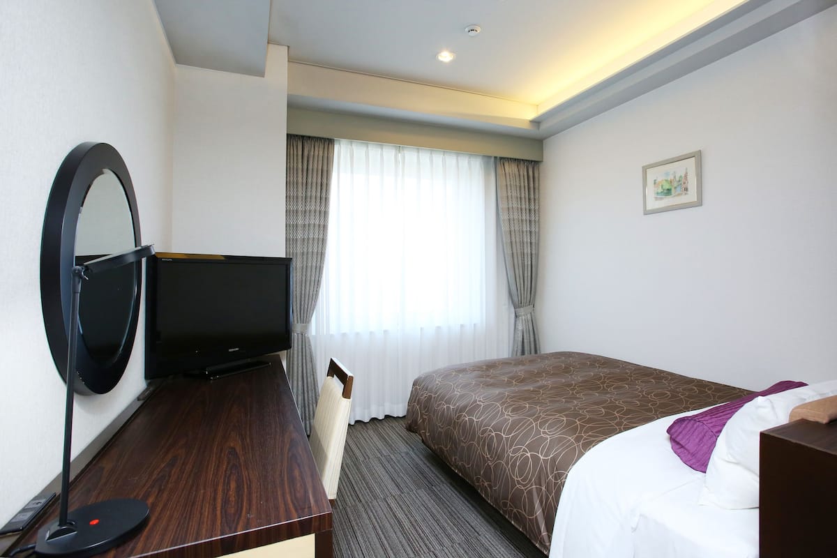 Habitación de hotel con cama, escritorio con computadora, televisión y ventana grande con cortinas.