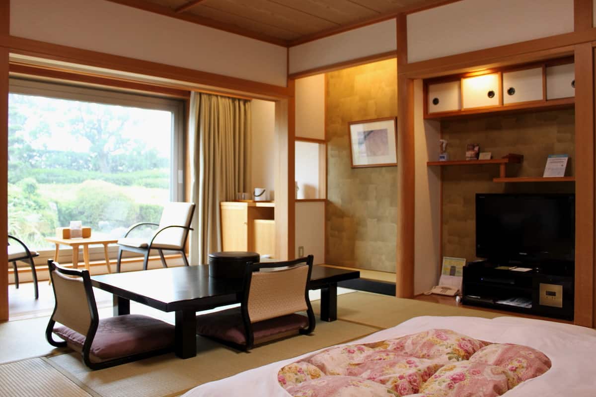Habitación de hotel con suelo de tatami, cama con ropa de cama floral, zona de comedor con mesa y sillas, televisor y vista a zonas verdes.