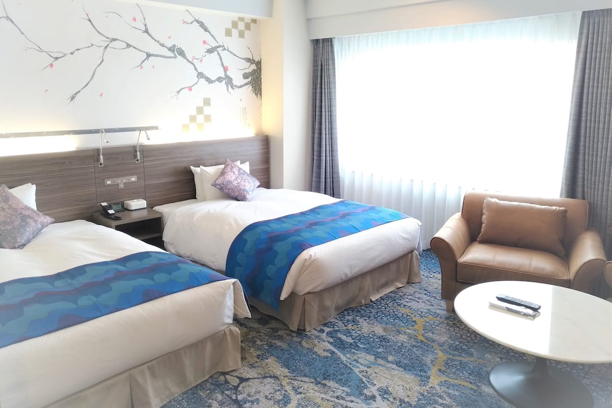 Habitación de hotel con dos camas, un sofá, una mesita y un ventanal grande con cortinas.