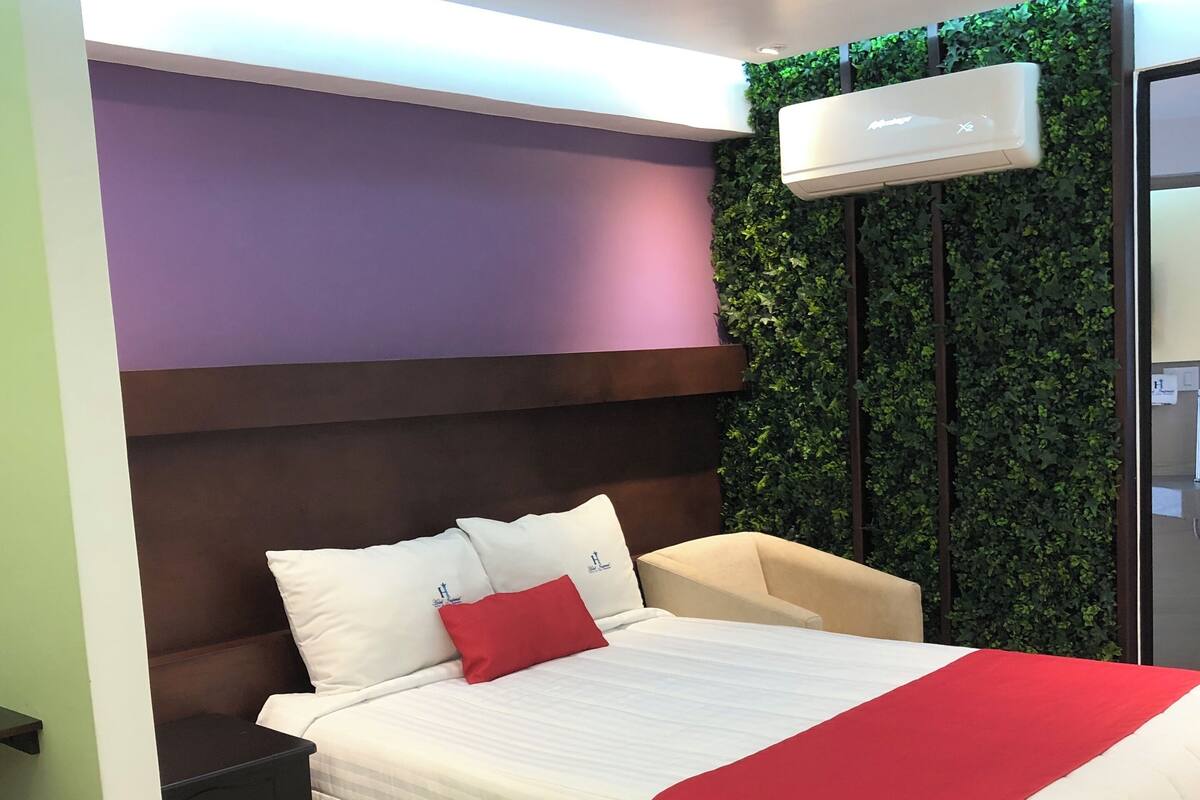 A modern bedroom with a bed, bedside tables, a chair, a wall-mounted light, and a green leafy wall.