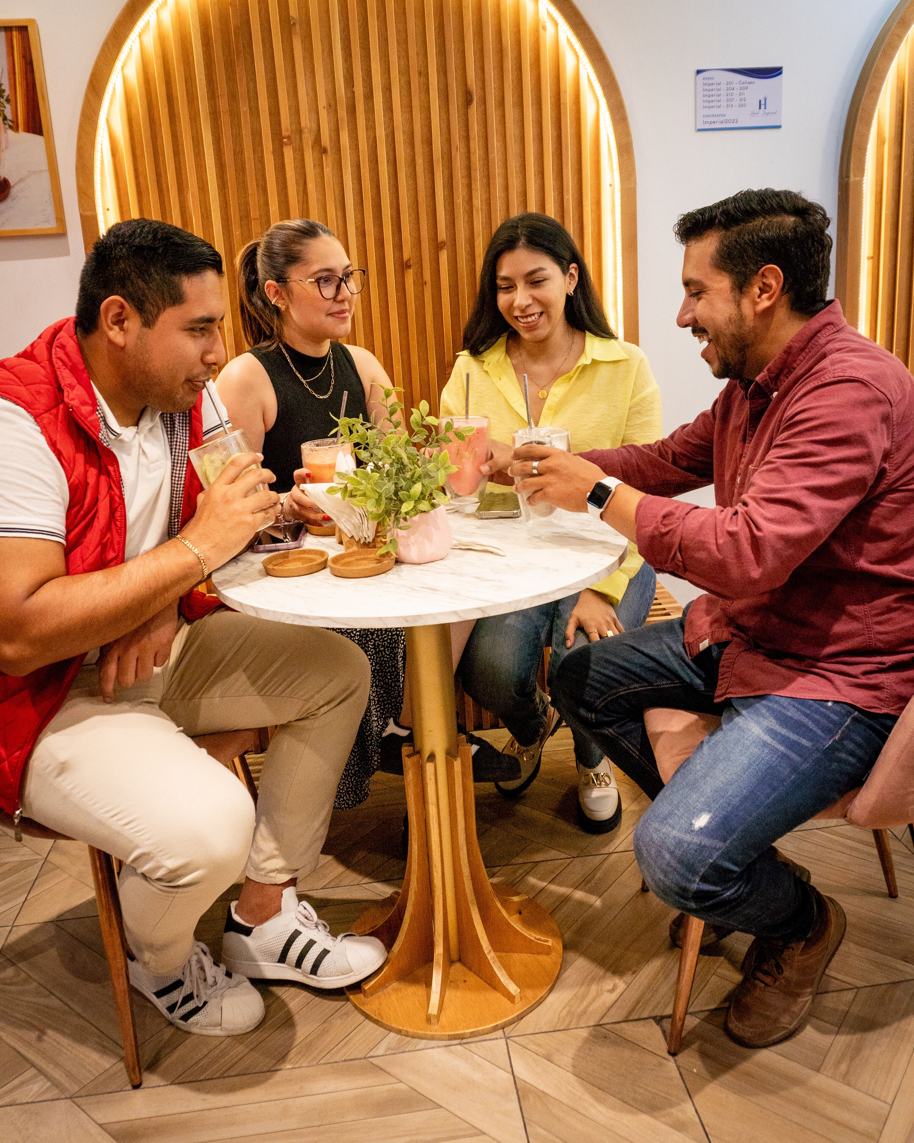 A group of people seated around a round table with drinks, in a room with wooden paneling and a tiled floor.