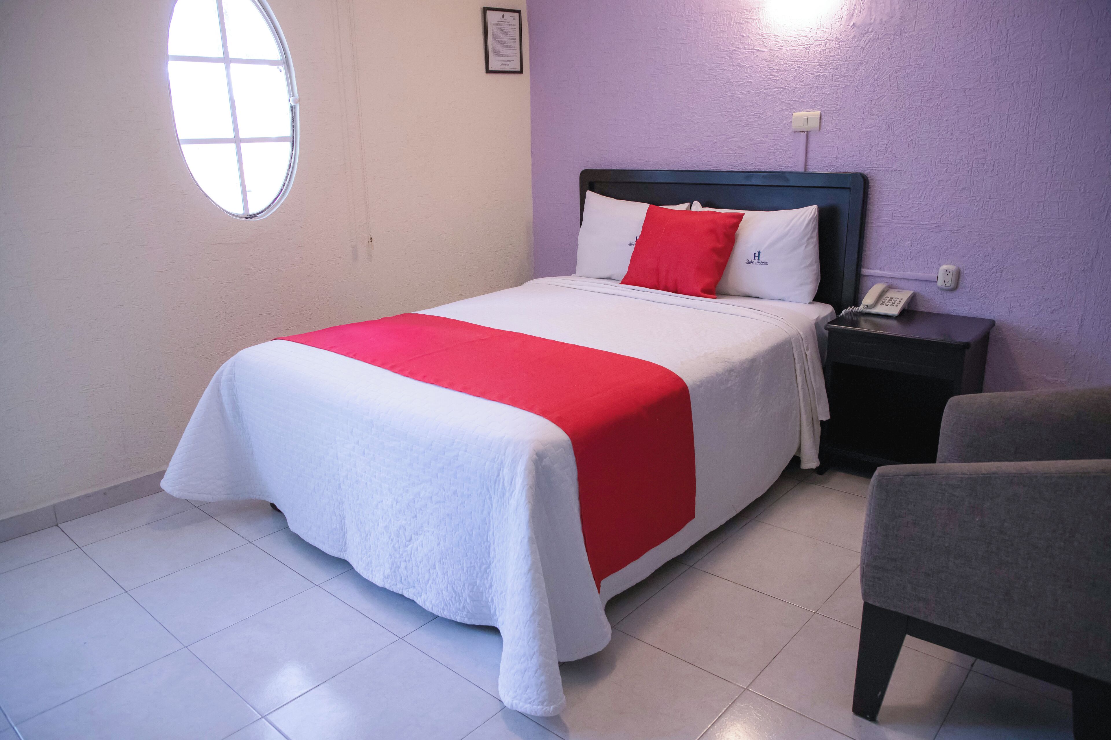 Habitación de hotel con cama, mesita de noche, silla, ventana y teléfono en la pared.