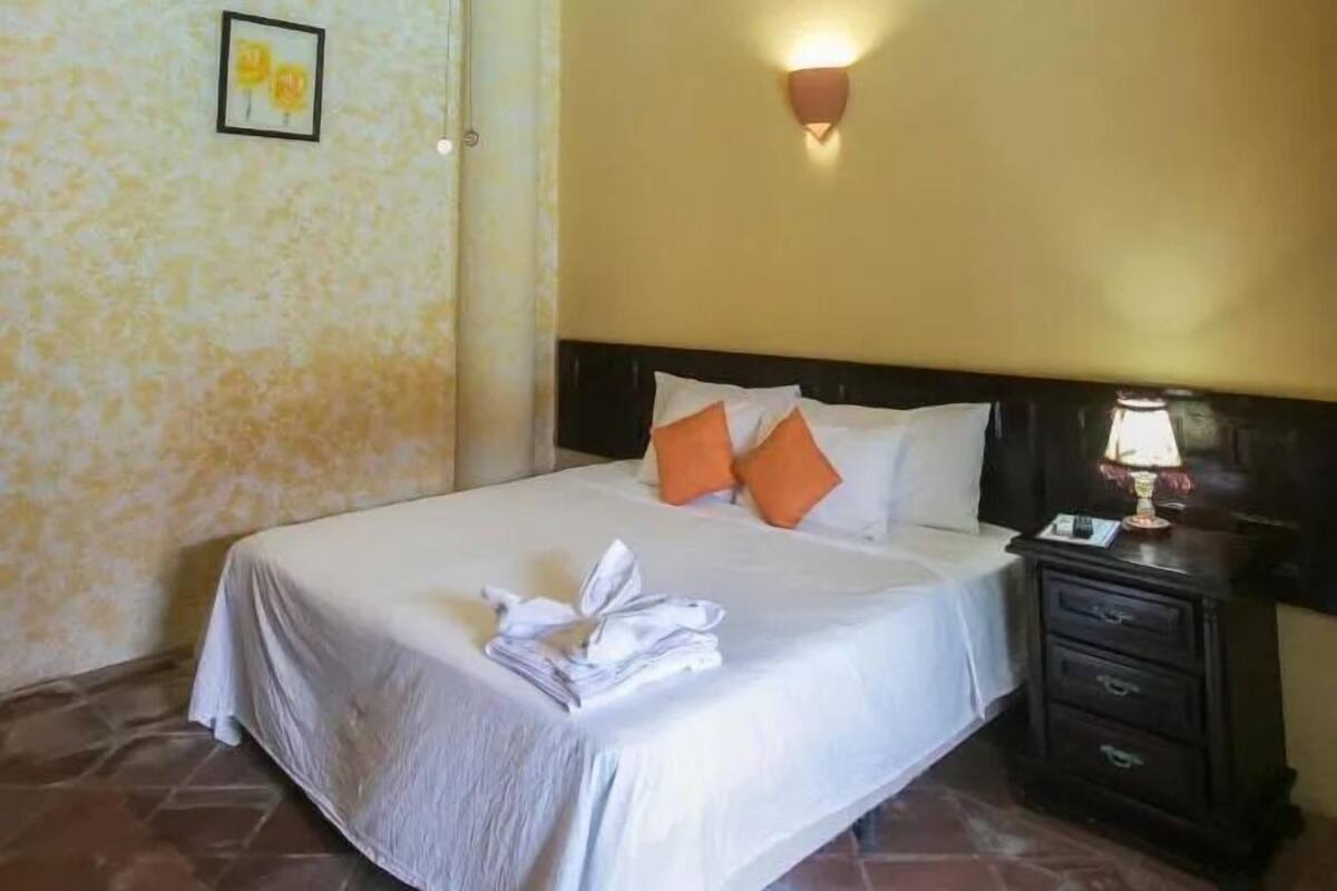 A neatly made bed with white linens and orange pillows, a bedside lamp, and a small framed picture on the wall.