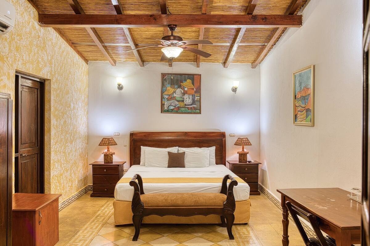 A bedroom with a wooden bed, two bedside tables, a bench, a ceiling fan, and artwork on the walls.