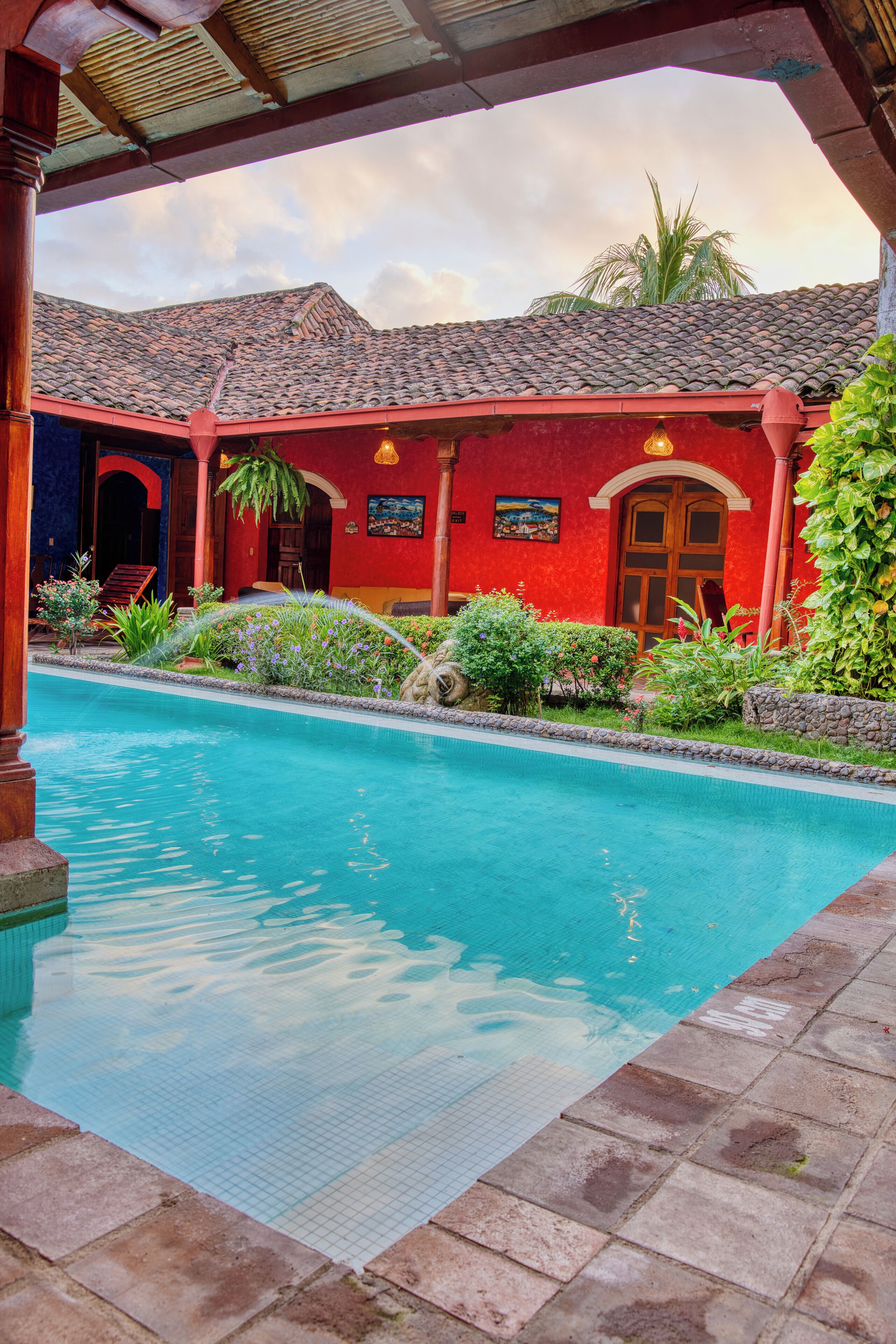 Una piscina con borde de azulejos, rodeada por un edificio rojo con techo de tejas y un área cubierta con vigas de madera.