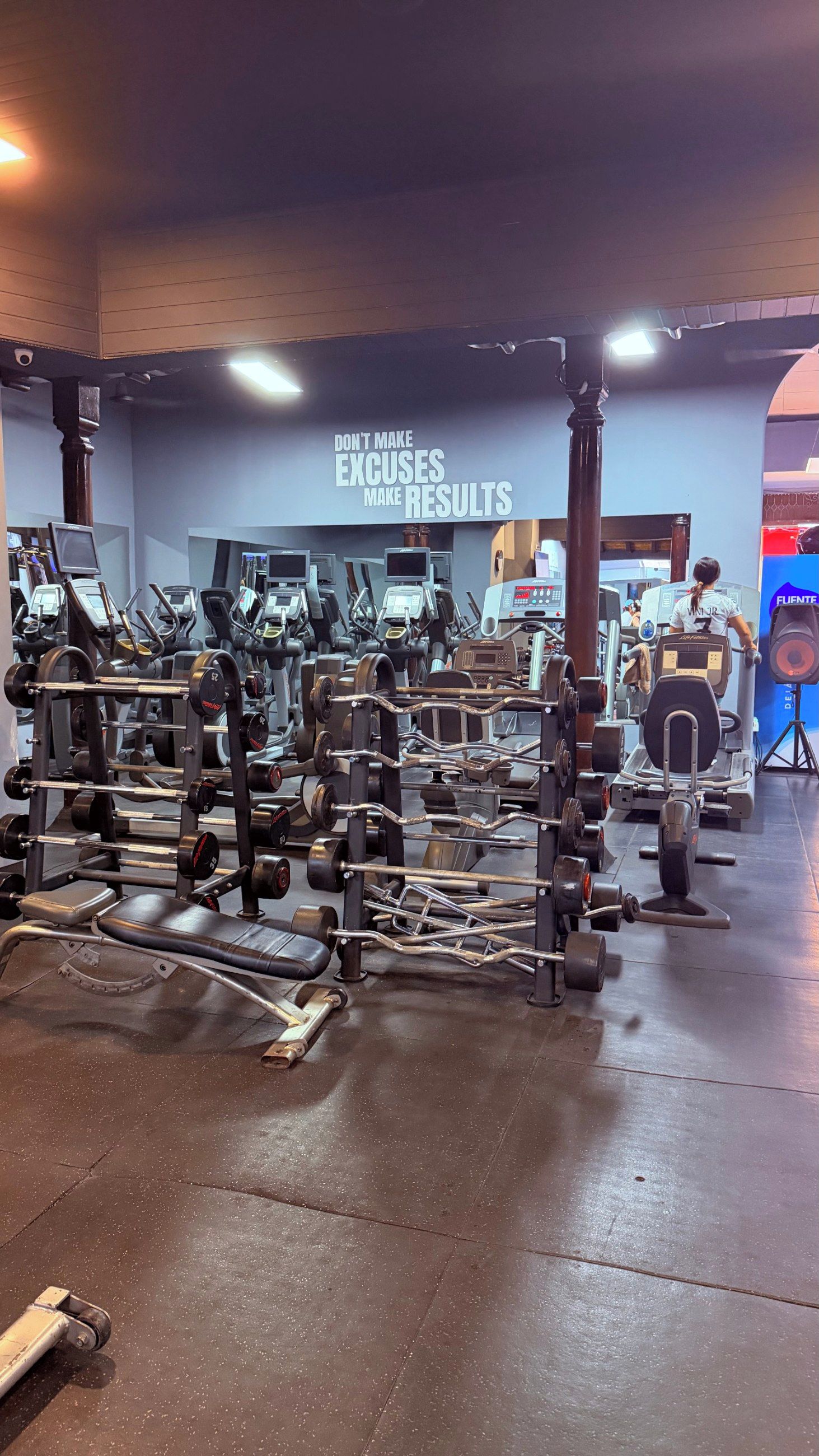 Une salle de sport équipée de divers appareils d’exercice, une citation motivante affichée sur le mur et une personne qui utilise un tapis de course.